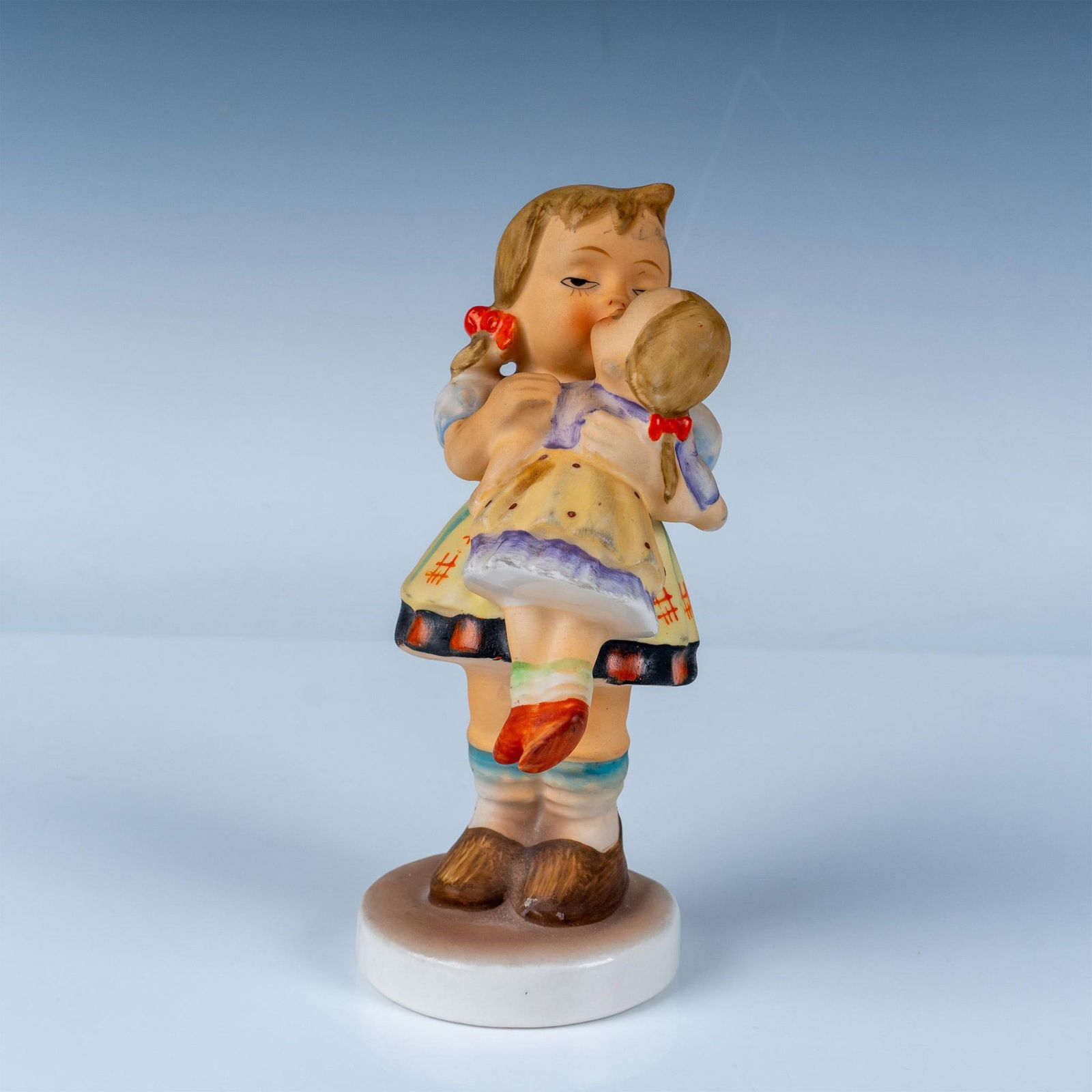 Hummel Goebel Porcelain Figurine Kiss Me Girl with Doll: This charming Goebel Hummel figurine titled Kiss Me depicts a young girl lovingly holding a doll close, sharing a tender moment. Crafted with precise attention to detail, this hand-painted porcelain p
