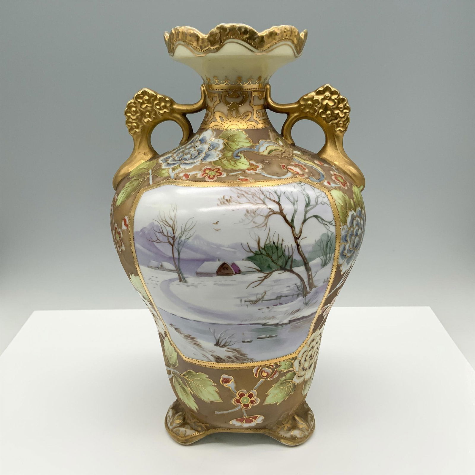 Nippon Footed Vase with Gilded Handles: Elegant hand painted ceramic vase featuring intricate floral designs in blue, green, and red hues. The vase includes ornate raised detailing, accented with a delicate gold finish on the handles and ri