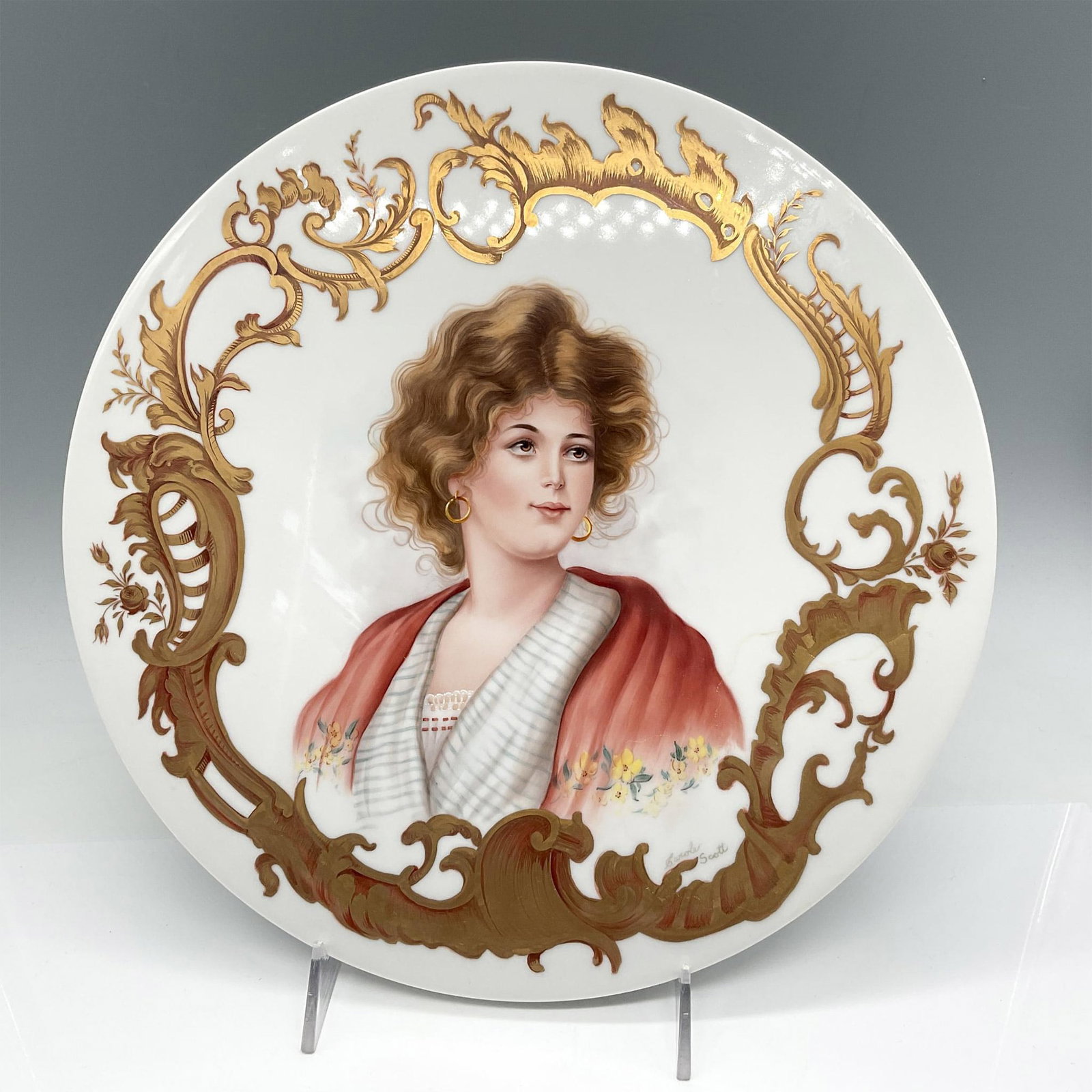 Carole Scott Porcelain Bust Plate, Signed: Elegant hand-painted portrait of a lady, adorned in a red shawl and white gown, artistically framed within elaborate gilt scrollwork. Signed by artist. Artist: Carole Scott Issued: 20th century Dimens