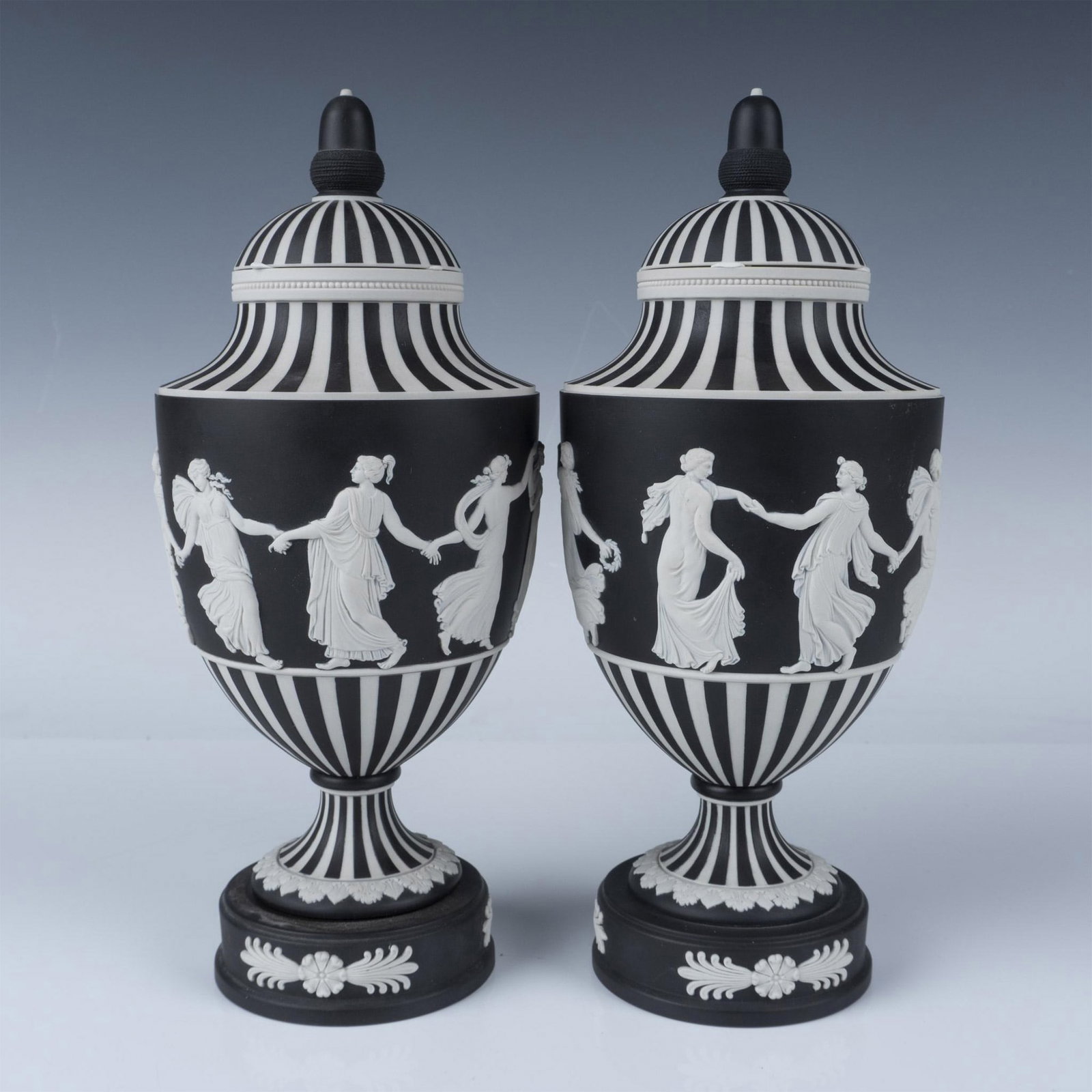 Pair of Wedgwood Black Basalt Jasperware Dip Covered Urns: Two white porcelain footed vases dipped in a black finish. Depicts, in white raised reliefs, maidens dancing. Includes original lid with an black acorn-shaped finial. Wedgwood Made In England impresse