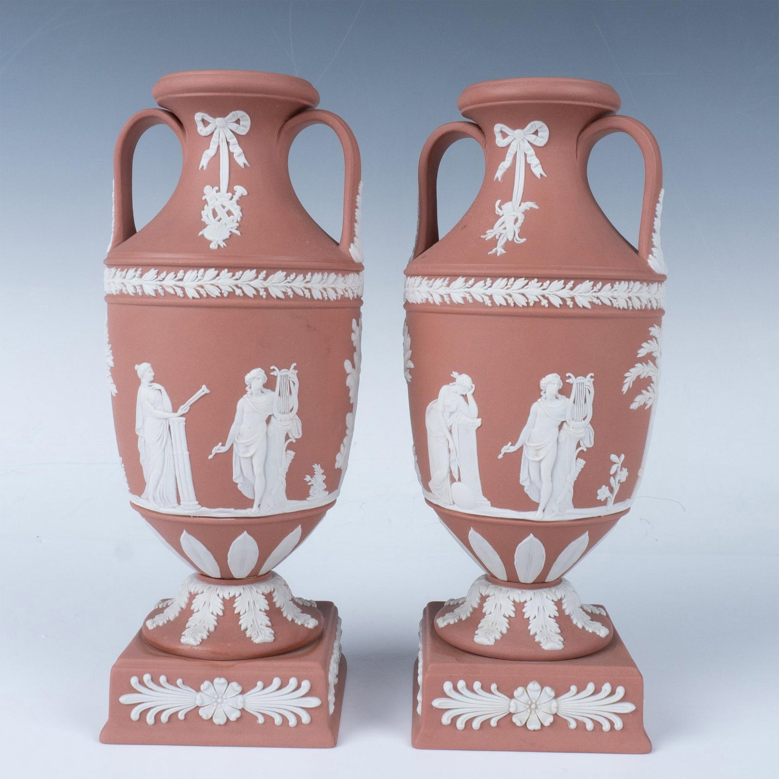 Pair of Wedgwood Terracotta Jasperware Urn Vases: A set of two urn-shaped terracotta vases attached to a square base. Each vase features a classical relief scene in white. Wedgwood, Made In England impressed stamp. Issued: 20th century Dimensions: 4"