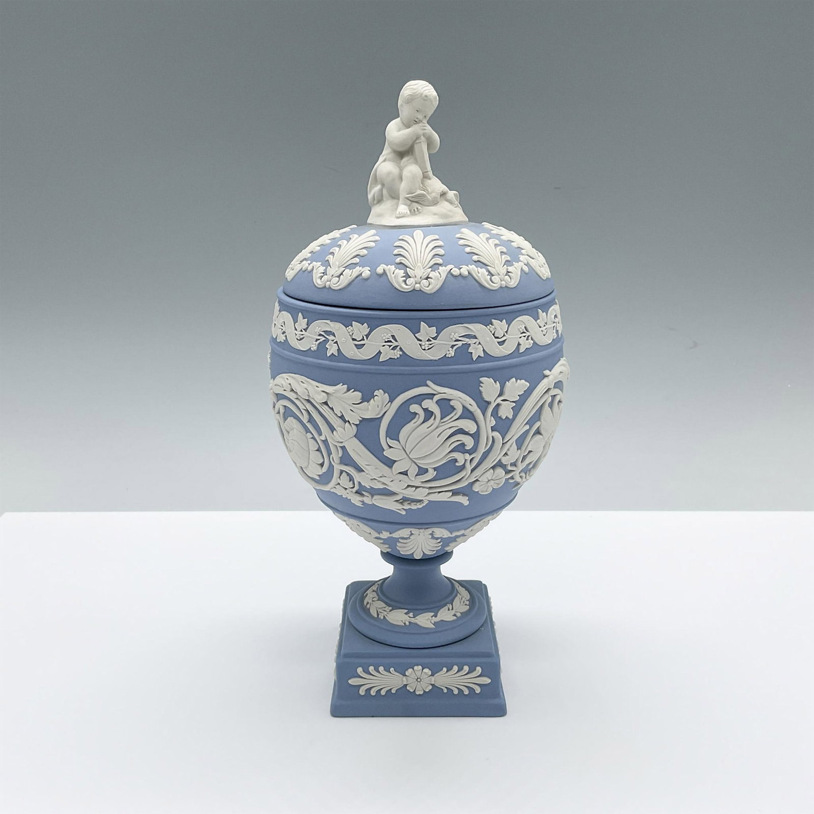 Wedgwood Pale Blue Jasperware Covered Vase: A beautiful blue urn-shaped vase with a round decorative lid and square base. The English pottery is adorned with floral blue reliefs. A putto seats atop of the lid as the finial. Wedgwood Made In Eng