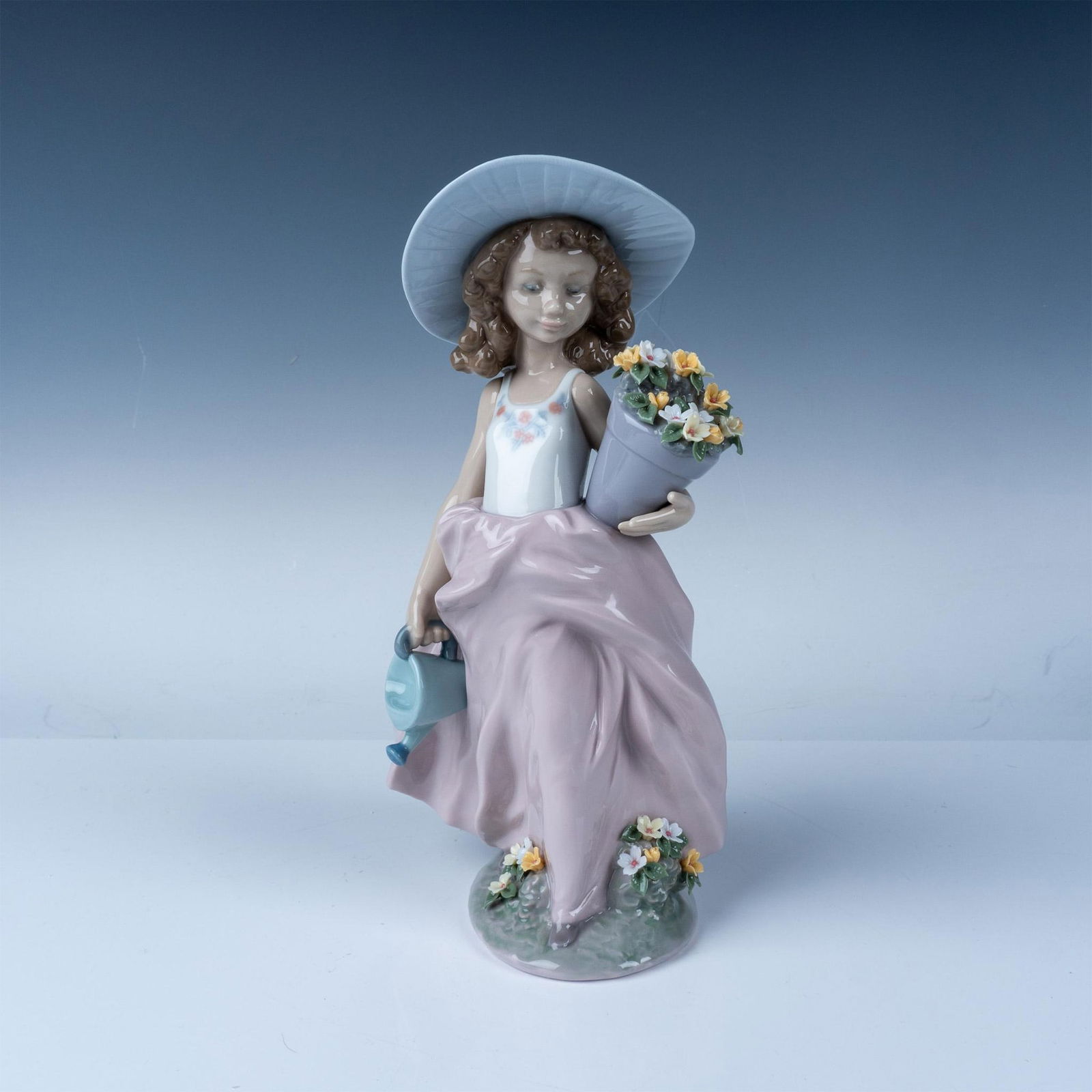 Lladro Figurine 07676 A Wish Come True with Original Box: This charming porcelain figurine by Lladro, titled Que bonitas!/ A Wish Come True, depicts a young girl elegantly dressed in a soft pink gown and sun hat, holding a bouquet of vibrant flowers and a wa
