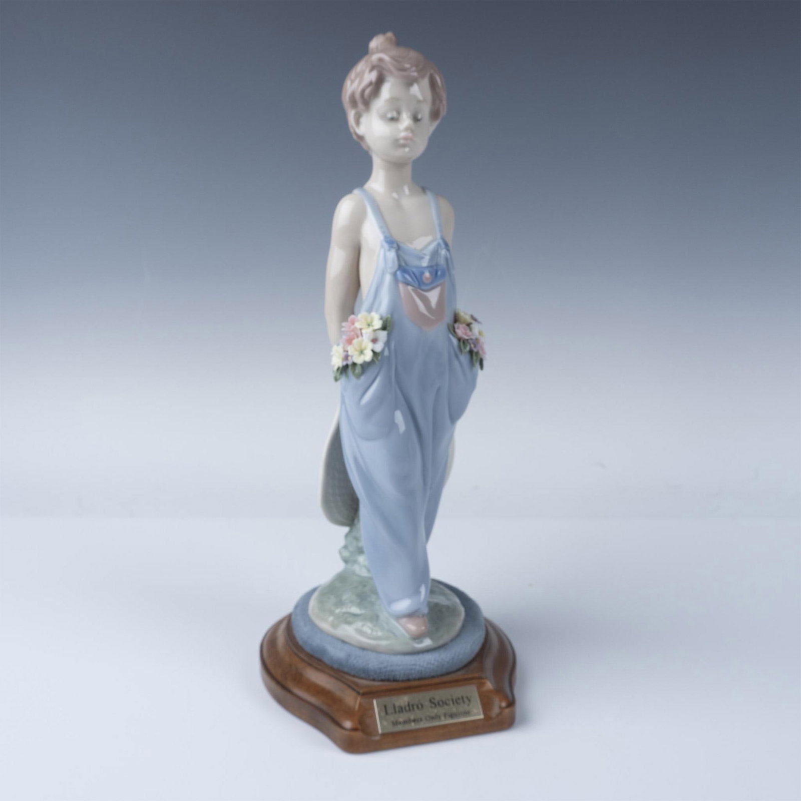 Lladro Pocket Full of Wishes Figurine No. 1007650 plus Base: A charming Lladro porcelain figurine titled Pocket Full of Wishes, depicting a young boy in blue suspenders with delicate floral accents in his pockets and holding a straw hat behind his back. This fi