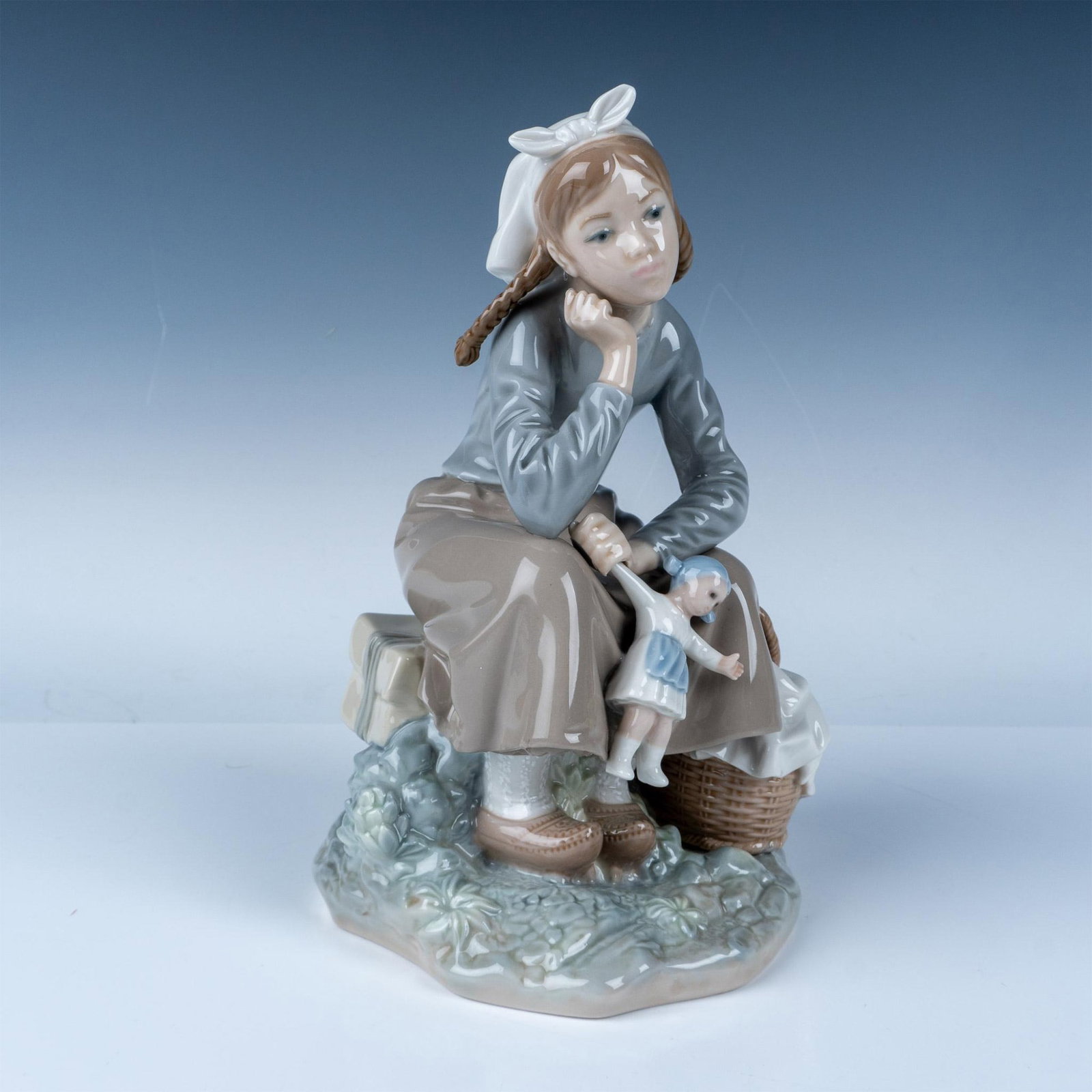 Lladro Porcelain Figurine Girl with Doll 1001211: Lladro porcelain figurine, number 1001211, featuring a seated young girl with pigtails holding a small doll while resting beside a basket of linens. The figurine reflects Lladro's craftsmanship in del