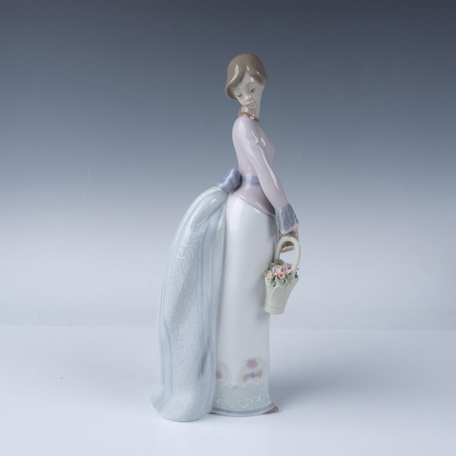 Lladro Porcelain Figurine Basket of Love 7622: Lladro Basket of Love figurine, model number 07622, portrays a graceful young woman delicately holding a basket of pastel flowers while elegantly gazing downward. Sculpted by Jose Puche and introduced