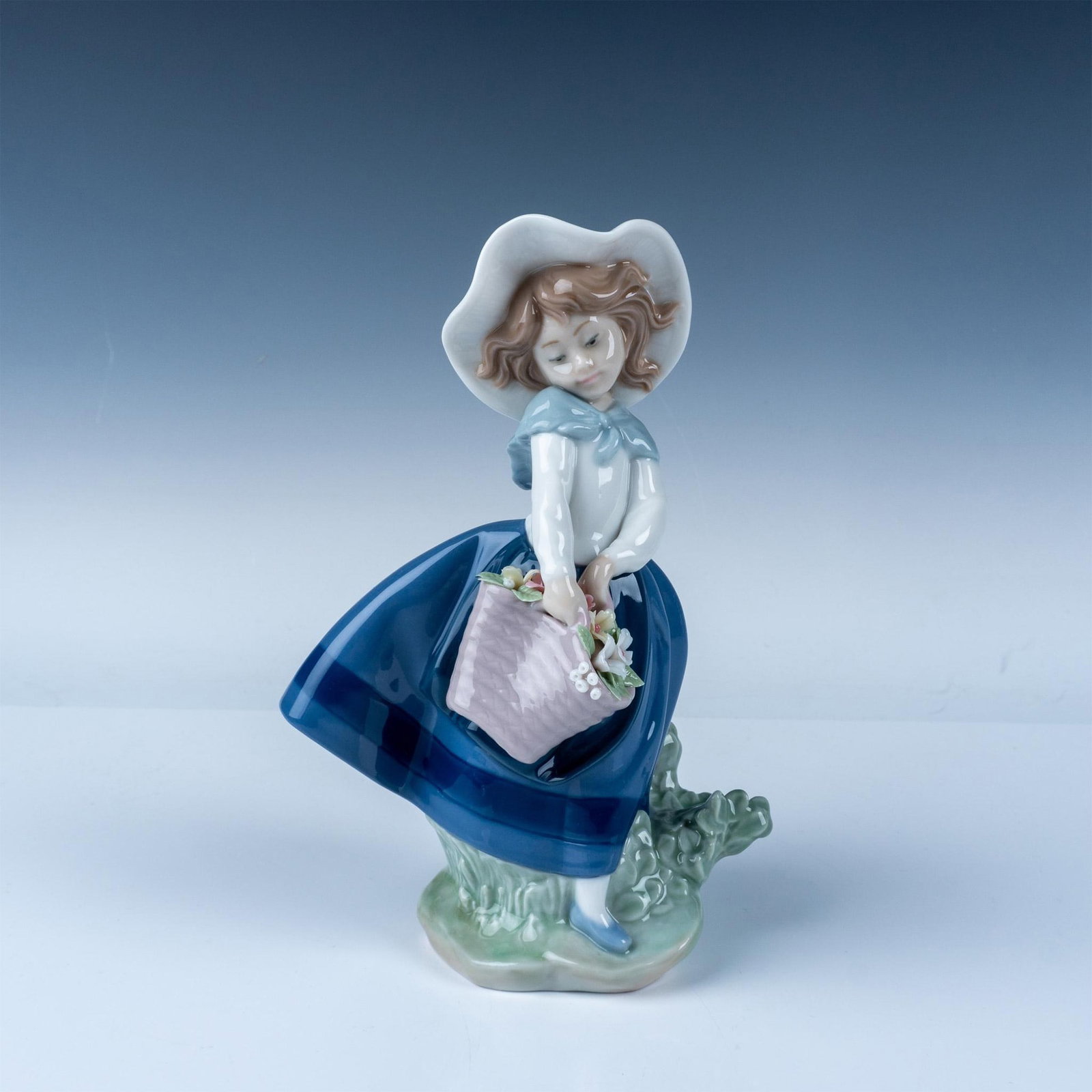 Lladro Porcelain Figurine Pretty Pickings No. 1005222: This exquisite Lladro porcelain figurine, titled Pretty Pickings, depicts a young girl adorned in a wide-brimmed hat and flowing blue skirt, gracefully holding a basket brimming with flowers. Sculpted