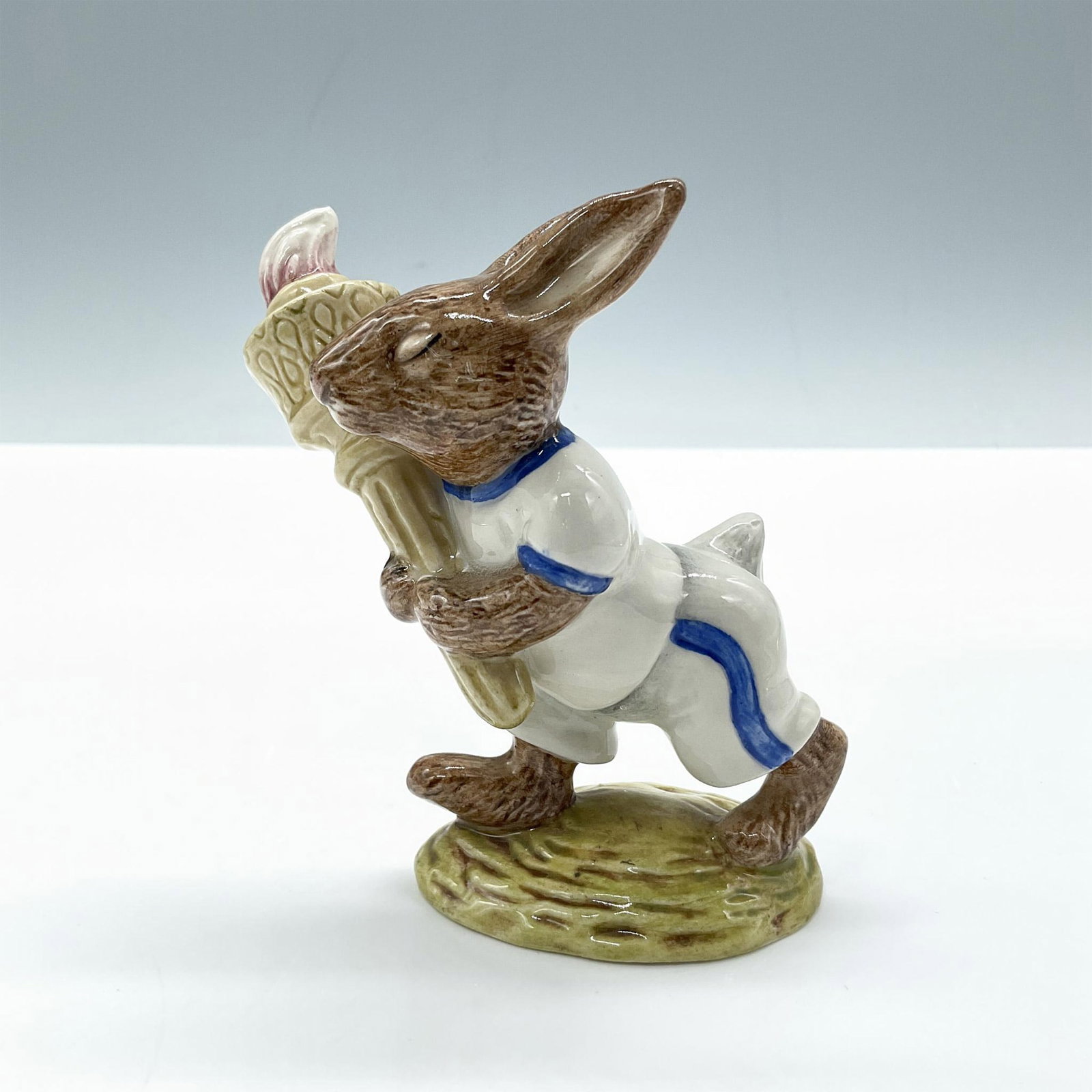 Olympic DB28 - Royal Doulton Bunnykins: A glossy porcelain figure of a bunny dressed in white and blue sports attire while holding a torch. Golden Jubilee Celebration 1984. Royal Doulton Bunnykins backstamp. Artist: H. Sales Issued: 1983 Di
