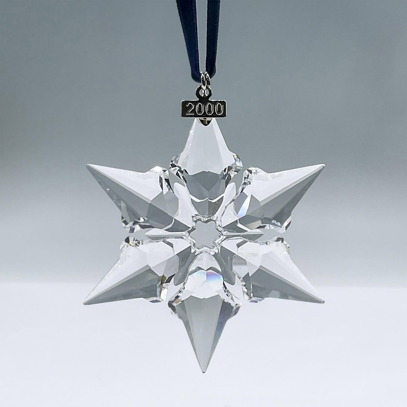 Swarovski Crystal Figurine, 2000 Christmas Ornament: This brilliant clear crystal ornament features 6 points, a metal tag with a 2000 engraving, and a blue satin ribbon. This item has its original box: 6.25"L x 1"W x 5.5"H. Swarovski acid mark. 243452. 