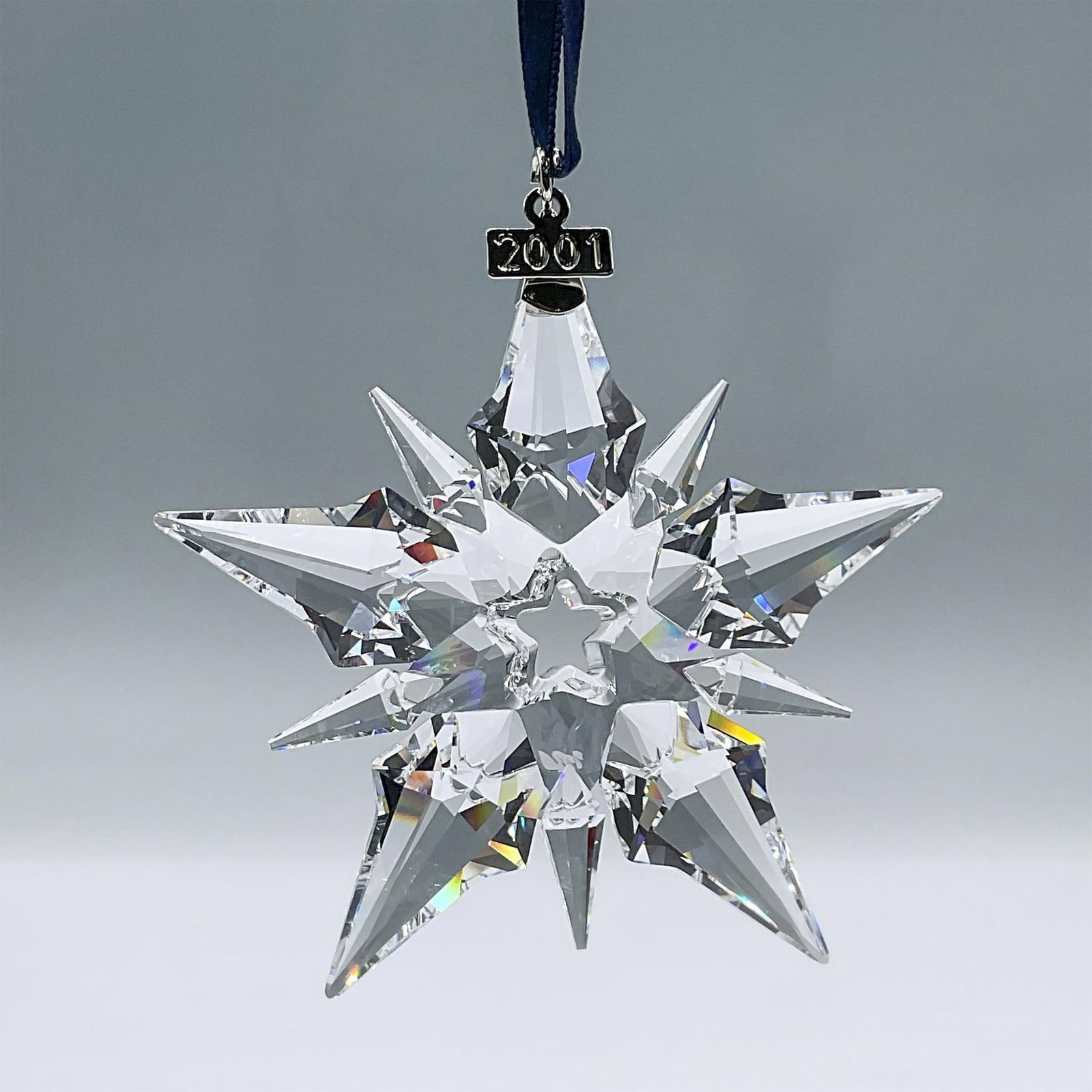 Swarovski Crystal Figurine, 2001 Christmas Ornament: This clear crystal ornament features 10 points, a metal tag with a 2001 engraving, and a blue satin ribbon. This item has its original box: 6.25"L x 1"W x 5.5"H. Swarovski acid mark. 267941. Artist: H
