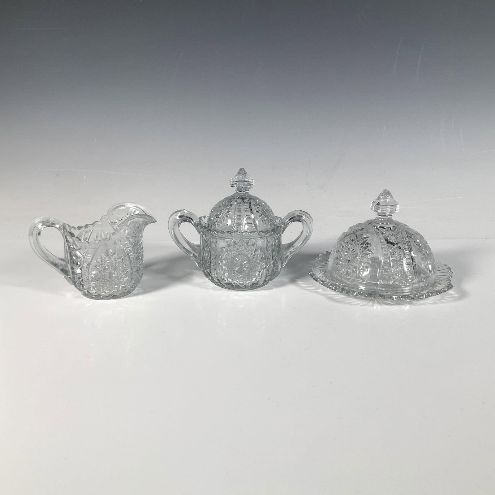 3pc Indiana Glass Oval Star Miniature Serveware: A stunning trio consisting of a butter plate with lid, a creamer, and a sugar bowl with lid, all featuring a star and geometric cut pattern. Butter plate with lid: 5.25"W x 3.5"H. Creamer: 4"L x 2.5"W