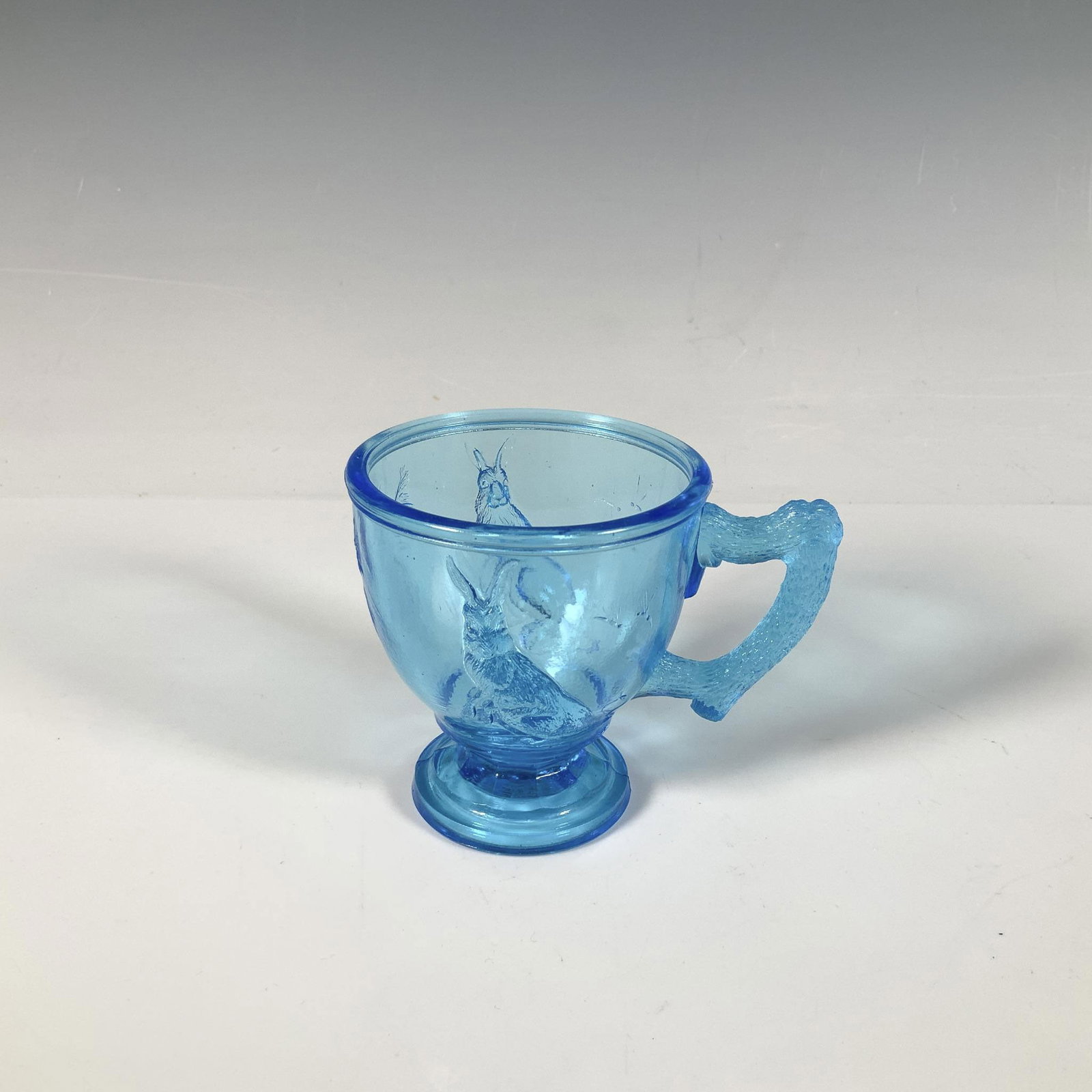 Blue Pressed Glass Rabbit Child's Mug: A charming small glass mug in a lovely blue hue featuring a high relief rabbit and wood-like handle. Dimensions: 4.25"L x 3"W x 3.25"H