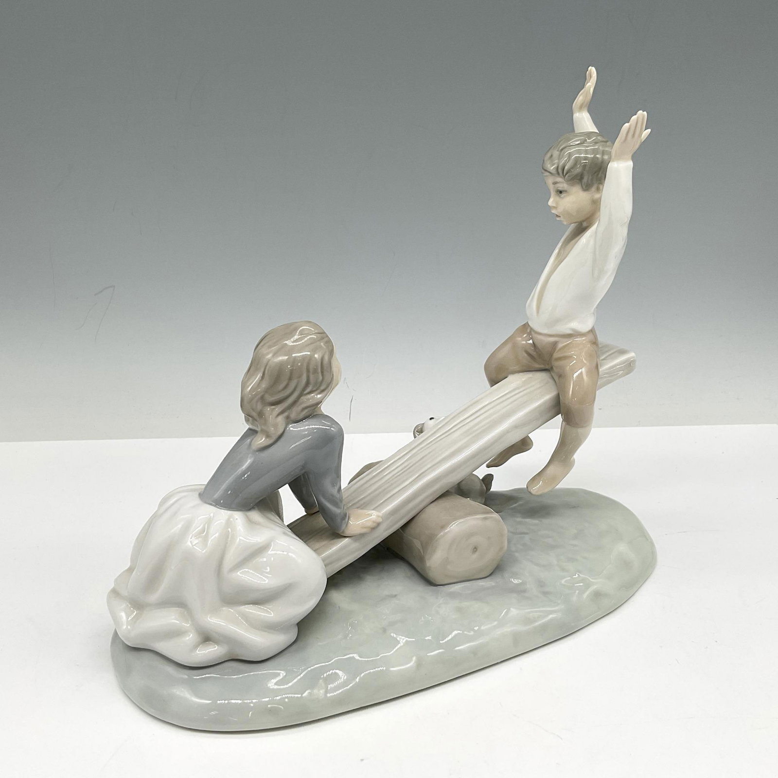 See-Saw 1004867 - Lladro Porcelain Figurine: Children playing on see-saw with dog looking on. Colored in cream, white and gray with glossy finish. This item has its original box: 12"L x 10"W x 6"H. Artist: Fulgencio Garcia Issued: 1974 - 1996 Di
