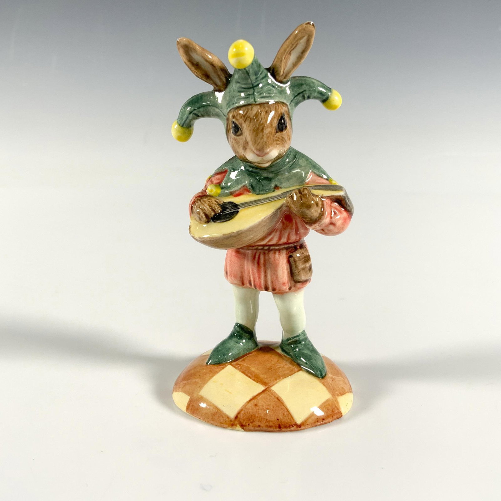 Jester DB161 - Royal Doulton Bunnykins: A whimsical bunny dressed in an orange and green jester outfit is poised to perform, holding a guitar and ready to entertain. Royal Doulton backstamp. Artist: D. Andrews Issued: 1995 Dimensions: 2"L x
