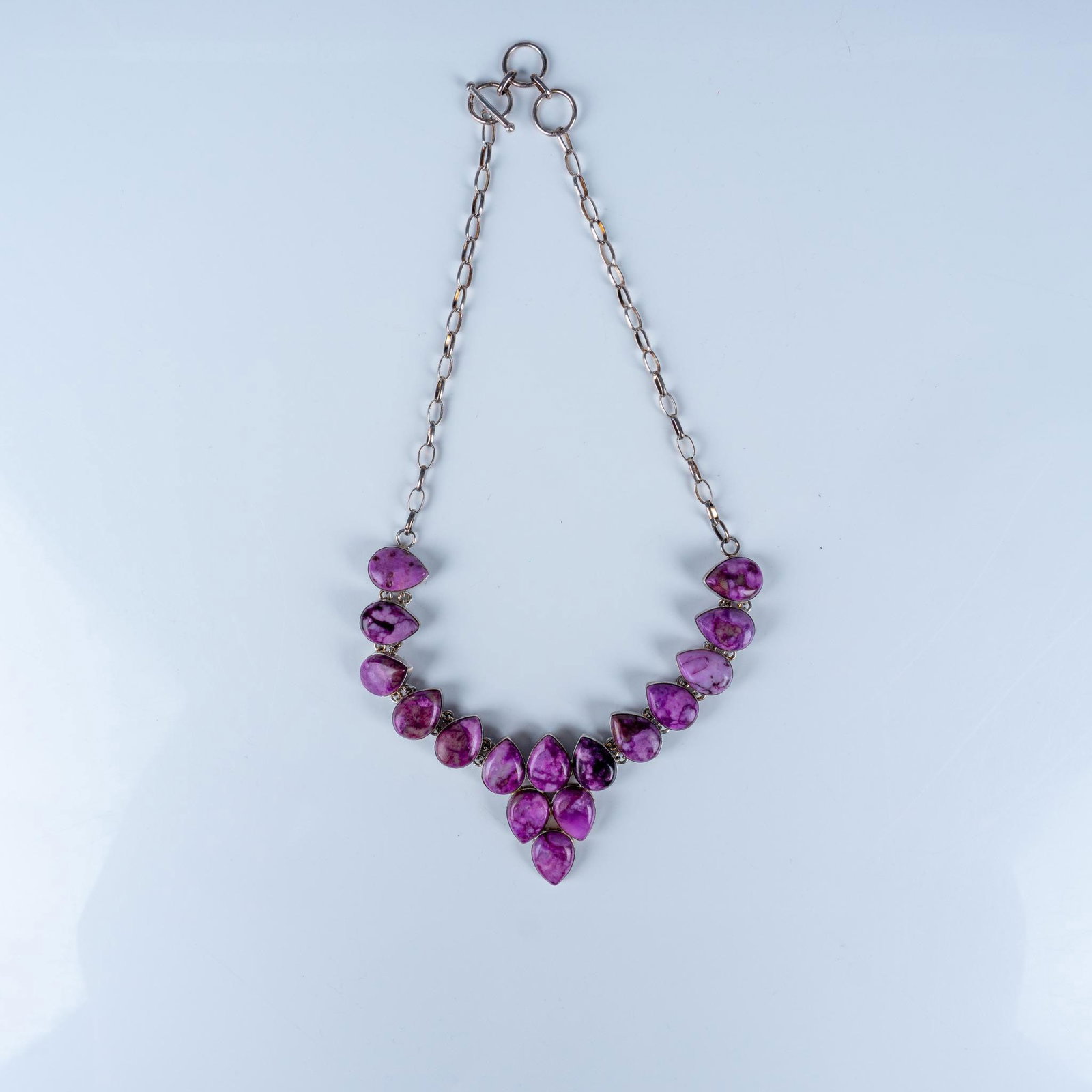 Silver and Purple Jasper Teardrop Necklace: A striking silver necklace featuring an arrangement of vibrant purple jasper stones cut into teardrop shapes. The stones showcase natural marbled patterns in shades of purple, pink, and white. The nec