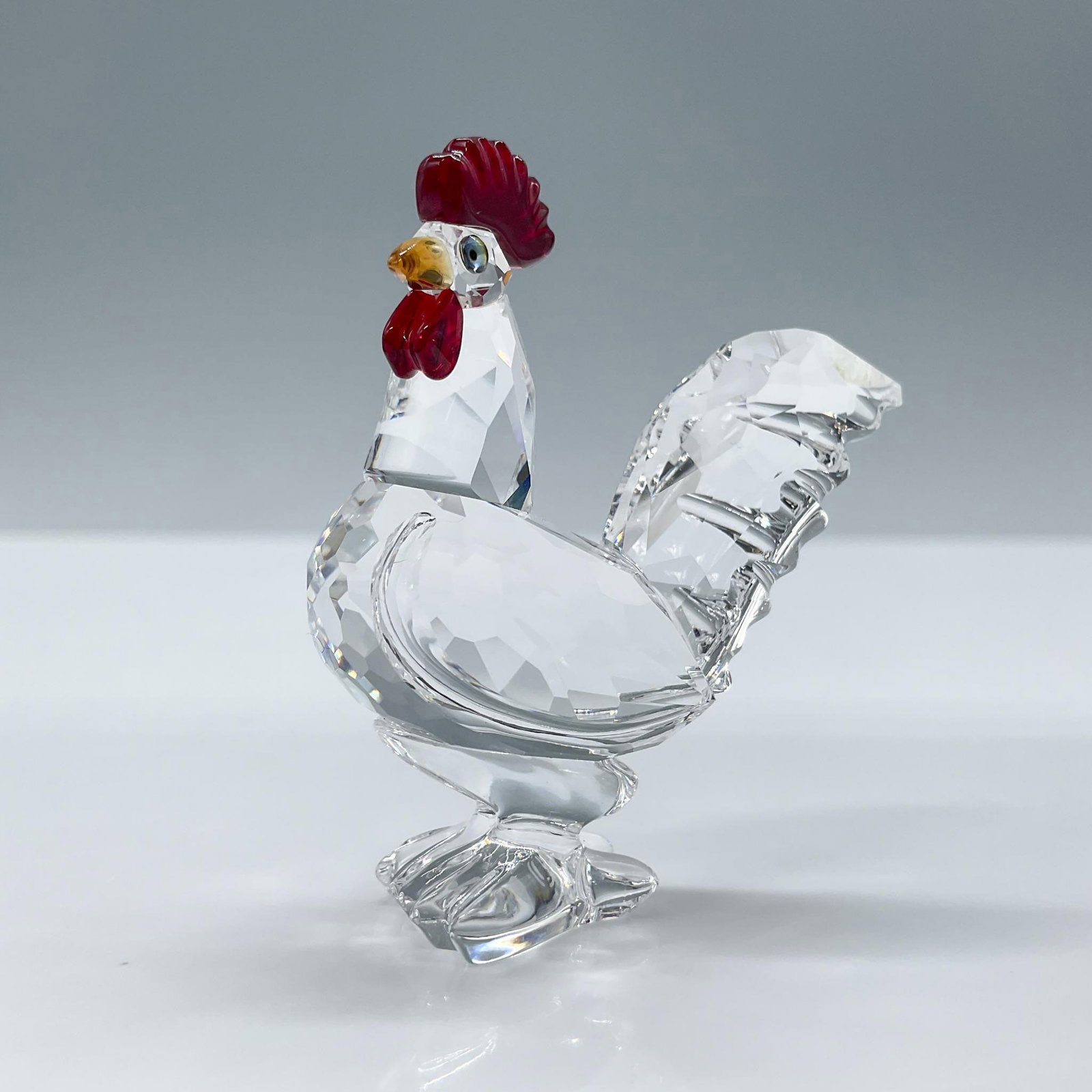 Swarovski Crystal Figurine, Cockrel with Color, 247759: Adorable clear crystal Cockrel with a red wattle, orange beak, and green gems for eyes. Swarovski etched backstamp. This item has its original box. Box measures 2.5"H x 3.25"dia. Artist: Michael Stame