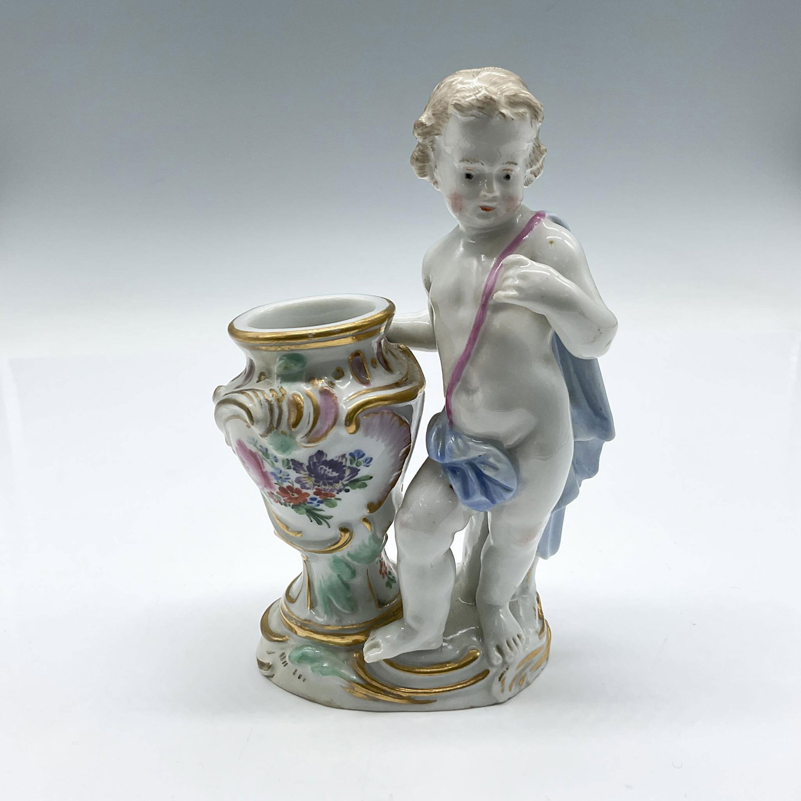 Meissen Porcelain Figurine, Cherub: Finely detailed porcelain figure is posed beside an elaborately decorated vase. The cherub is draped in light blue cloth with a pink sash and is in a naturalistic stance. Meissen mark. 957. Issued: 20