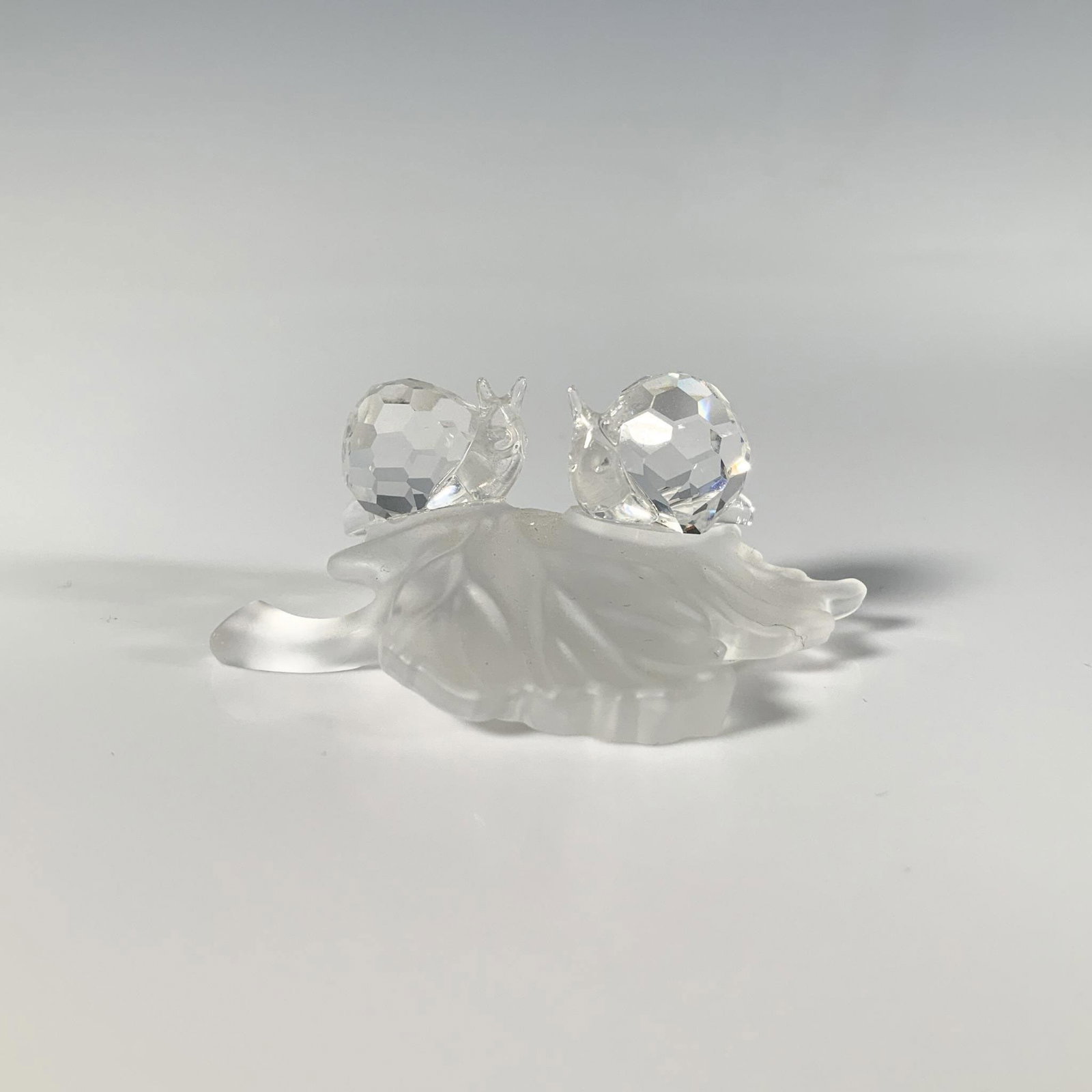 Swarovski Crystal Figurine, Baby Snails on Leaf: Two delightful precision-cut crystal snails perched on a smooth frosted leaf. Swarovski marking. This item has its original box: 3.25"W x 2.5"H. Artist: Edith Mair Issued: 2001-2009 Dimensions: 2"L x 