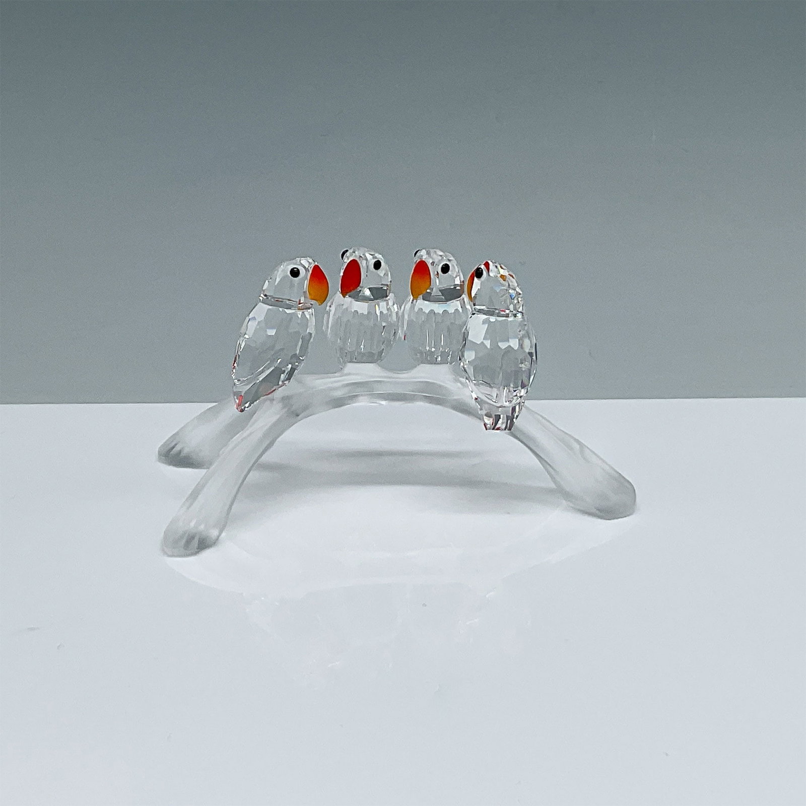 Swarovski Crystal Baby Lovebirds Figurine with Box: A Swarovski crystal figurine titled Baby Lovebirds, featuring four intricately crafted birds perched on a frosted crystal branch. The lovebirds showcase precision-cut clear crystal bodies with orange 