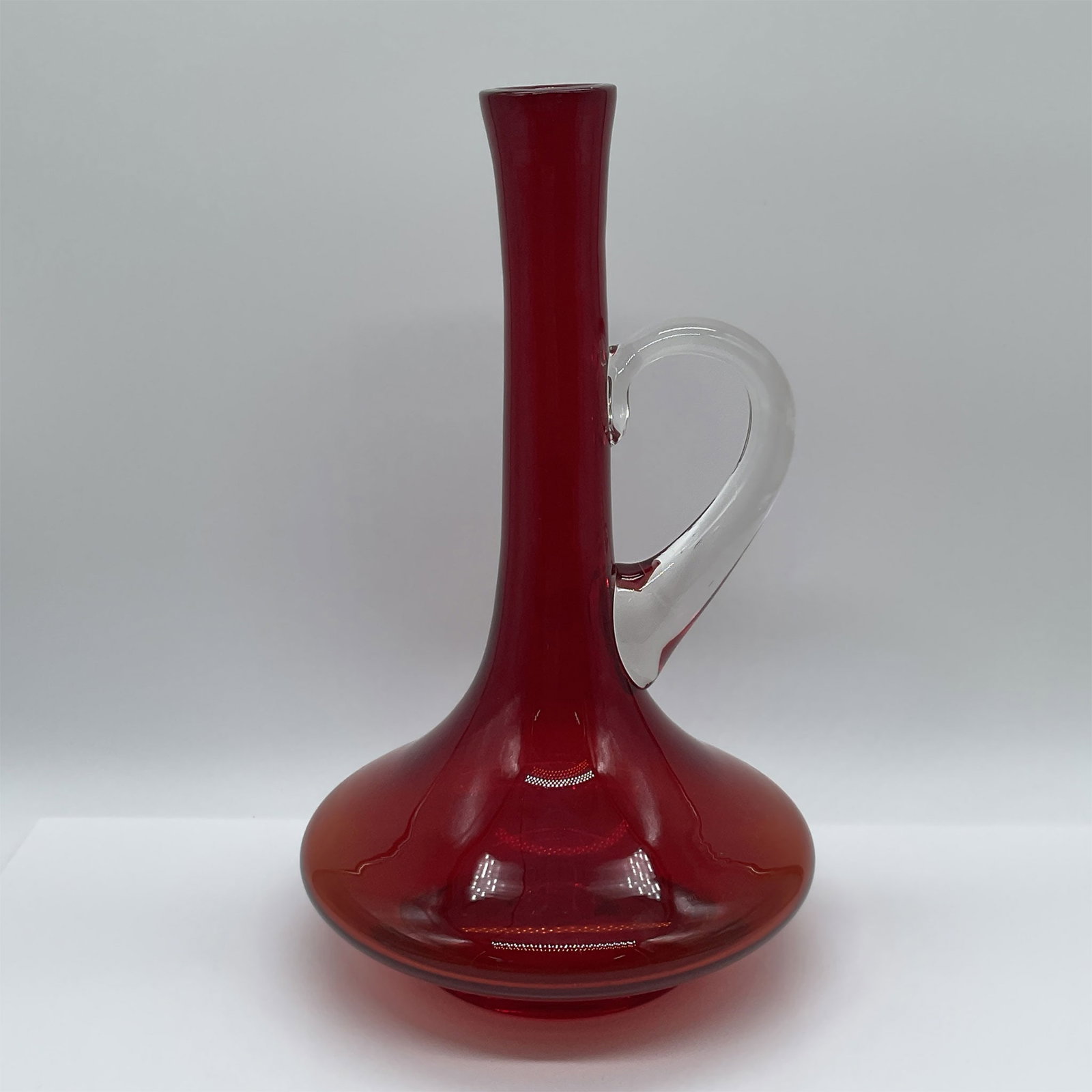 Mid-Century Red Glass Ewer: A striking mid-century glass ewer in vibrant red. The design features an elongated neck, wide base, and an elegantly arched clear glass handle, showcasing exceptional craftsmanship. This hand-blown pi