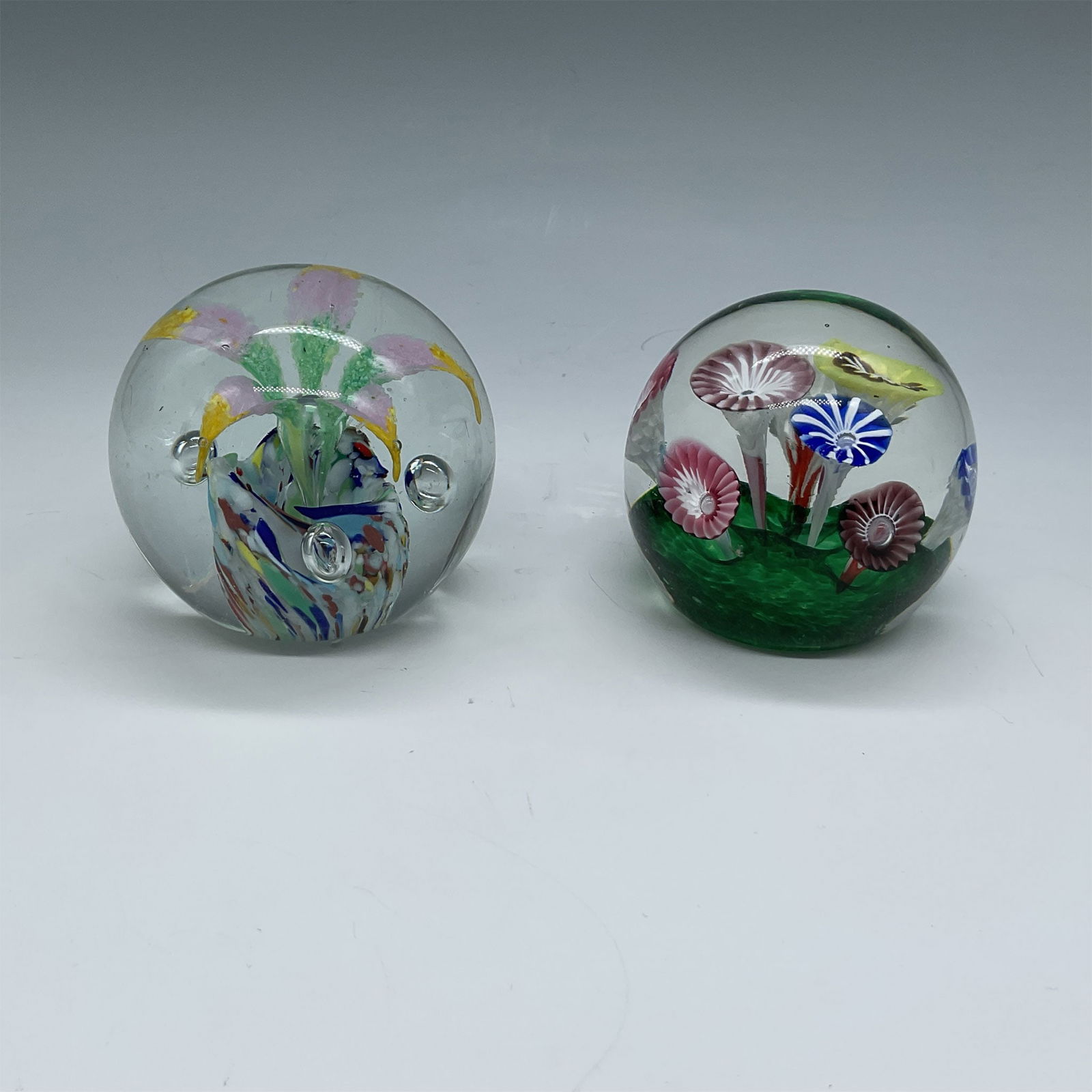 2pc Floral Design Paperweights: This lot features two paperweights with intricate floral designs. One paperweight showcases a vibrant pink flower with a multicolored base, while the other depicts a lush green field with multicolored