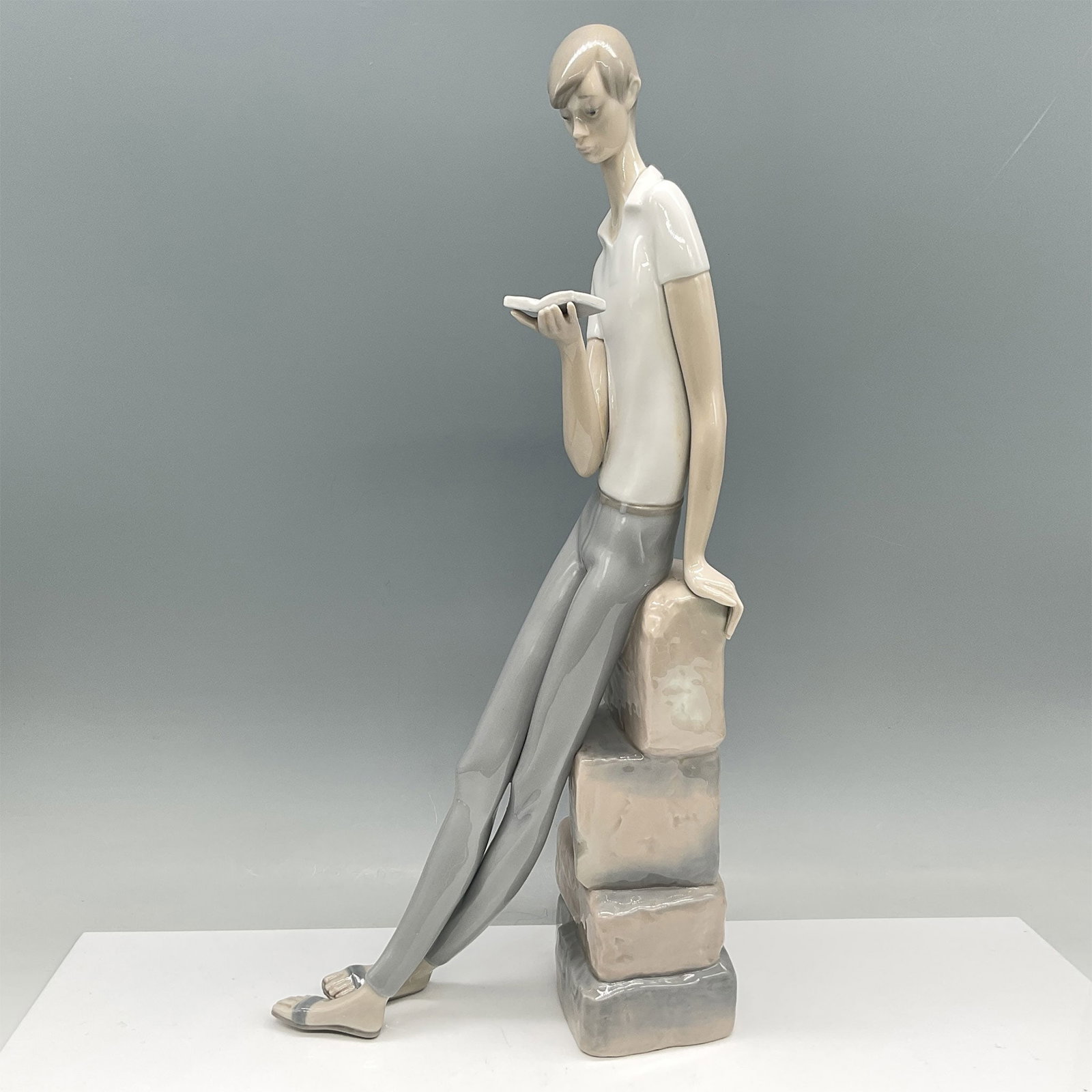 Lladro Porcelain Figurine, Boy Student 01004517: Charming Lladro porcelain figurine titled Boy Student, featuring a young man seated on a stack of stone blocks, deeply engrossed in a book. Designed by Fulgencio Garcia, this piece exemplifies Lladro 