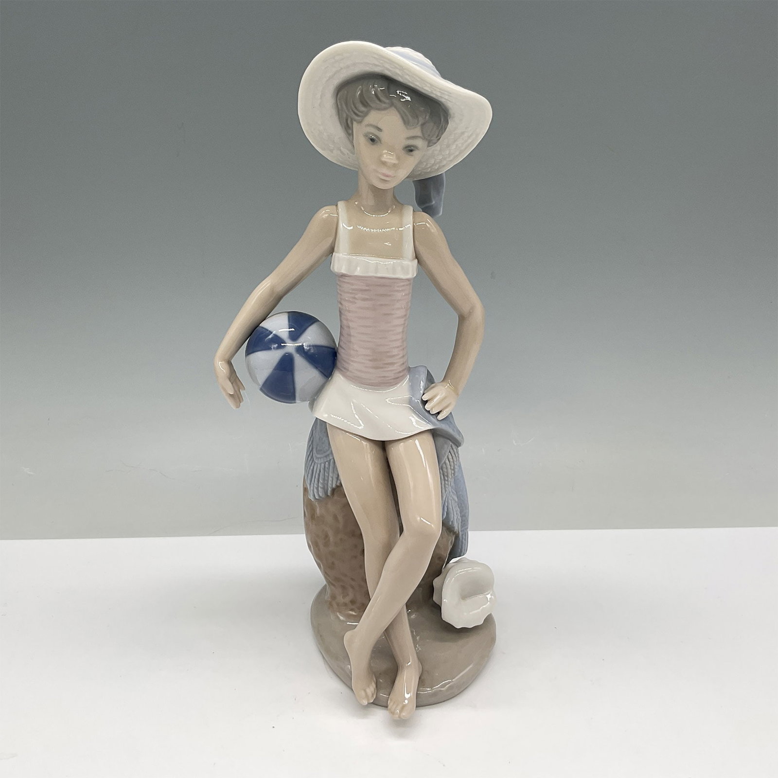 Lladro Porcelain Figurine, Summer 01005219: Charming Lladro porcelain figurine titled Summer, model number 01005219. Depicts a young girl in a relaxed pose, sitting with a beach ball and wearing a wide-brimmed hat. Expertly hand-painted in past