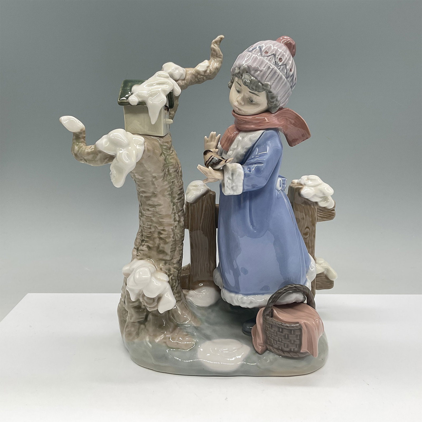 Lladro Porcelain Figurine, Winter Frost 01005287: This beautifully detailed Lladro porcelain figurine, titled Winter Frost, depicts a young girl in a winter setting. Dressed in a cozy blue coat, scarf, and hat, she interacts tenderly with a bird perc