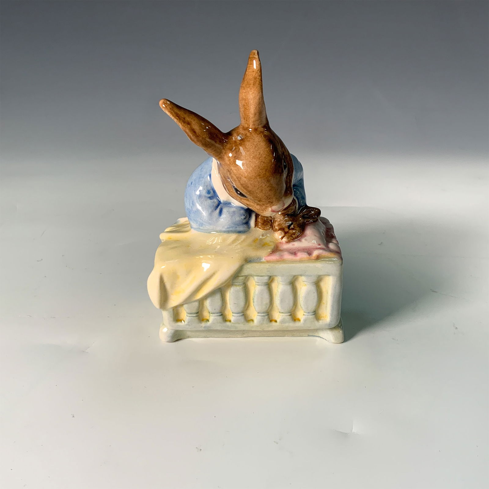Royal Doulton Bunnykins Figurine, New Baby DB158: This Royal Doulton Bunnykins figurine, titled "New Baby" (DB158), features a mother bunny wearing a blue dress with a white collar, lovingly rocking a cradle. The cradle is adorned with a pink pillow