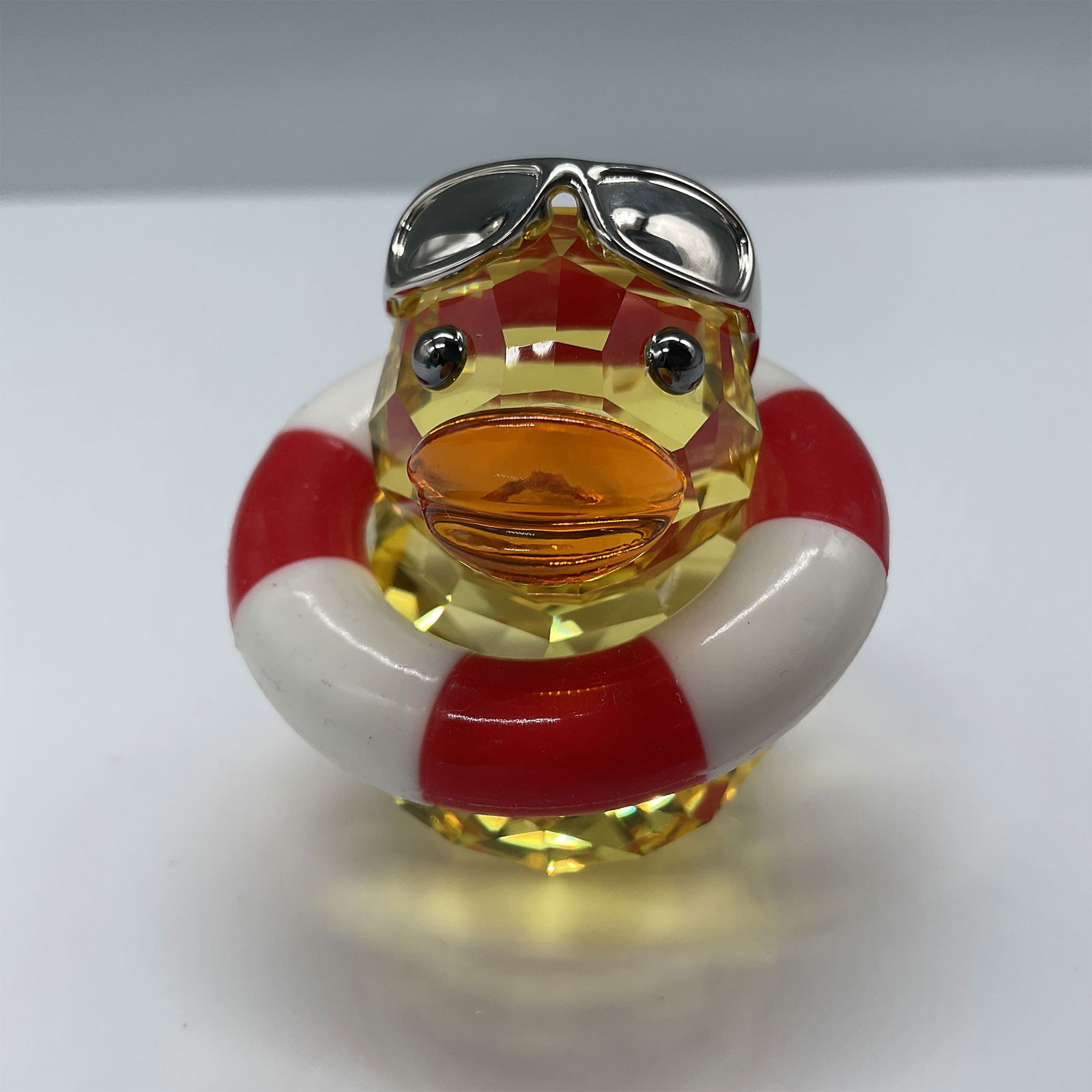 Swarovski Crystal Figurine, Sunny Sam with Box: Swarovski crystal figurine "Sunny Sam" from the Happy Ducks collection, measuring 2"H. The piece features vibrant colors with Sunny Sam donning sunglasses and a lifebuoy. Comes in its original box, me