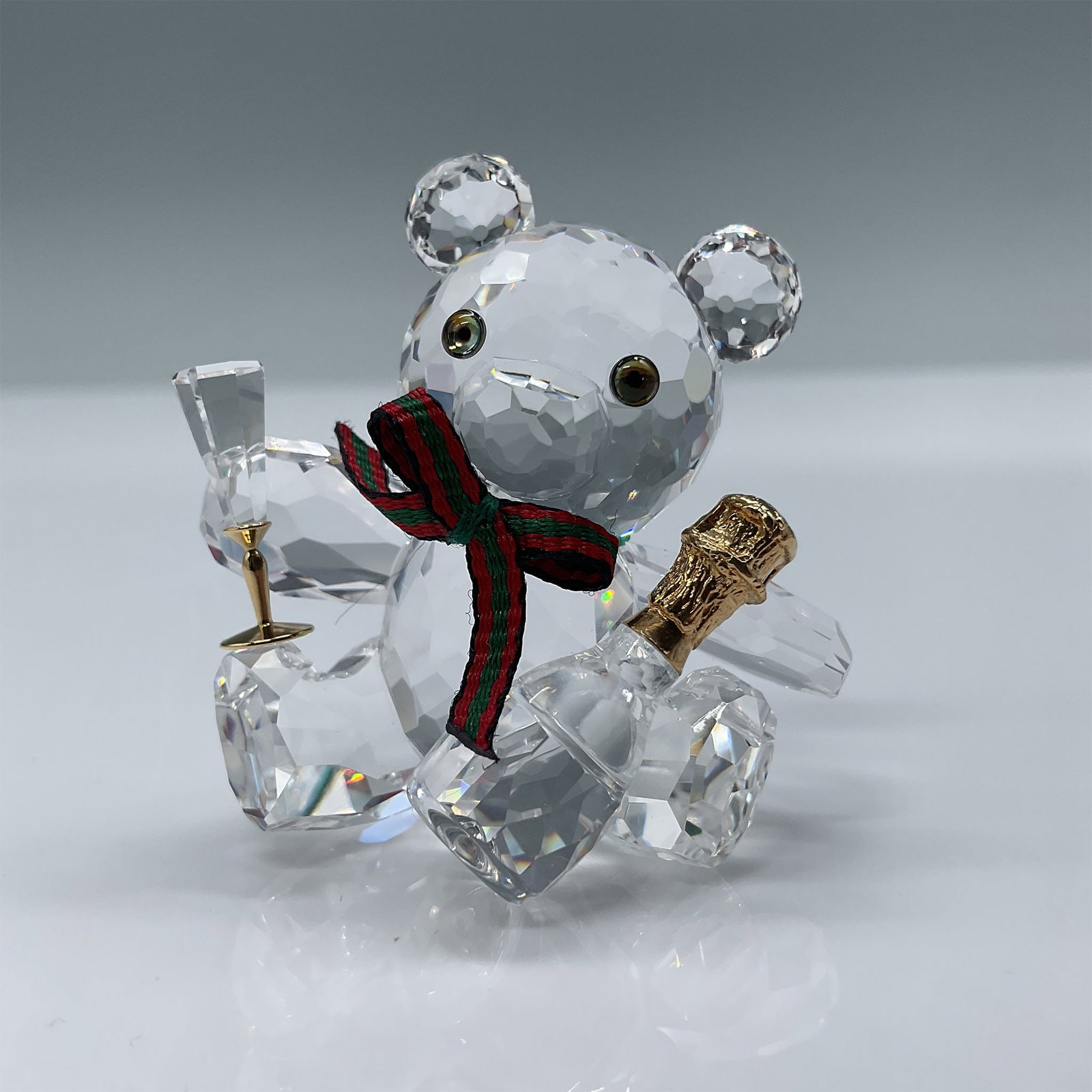 Swarovski Crystal Figurine, Kris Bear Champagne Toast: Swarovski Crystal figurine featuring Kris Bear celebrating with a champagne toast. This charming design includes intricate faceted crystal details, a red scarf, and golden champagne bottle accents. Th
