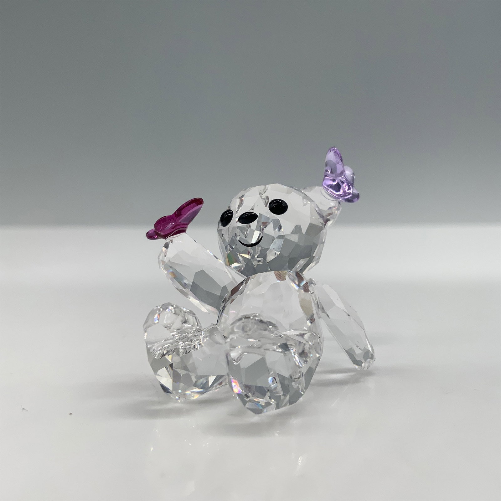 Swarovski Kris Bear Playful Butterflies Figurine with Box: Introducing the delightful Swarovski Kris Bear Playful Butterflies figurine, a charming addition to any collection. This adorable piece features a clear crystal bear with a printed black smile, adorne