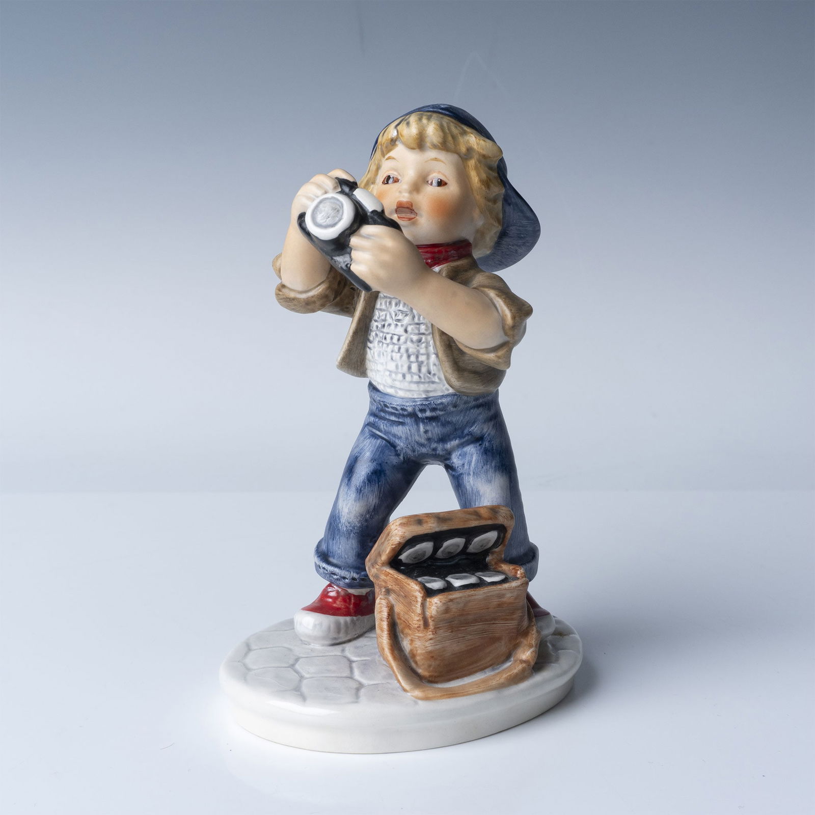Goebel Porcelain Figurine, Young Photographer: Goebel porcelain figurine titled "Young Photographer," featuring a young boy enthusiastically holding a camera with a bag of film rolls at his feet. Marked "Goebel W. Germany" and dated 1978 on the un