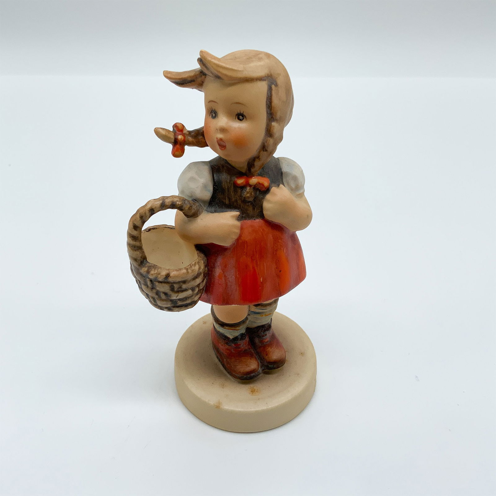 Goebel Hummel Porcelain Figurine, Little Shopper: This charming Hummel porcelain figurine, titled "Little Shopper," portrays a young girl holding a basket, dressed in a traditional outfit with a red skirt, white blouse, and braided hair adorned with