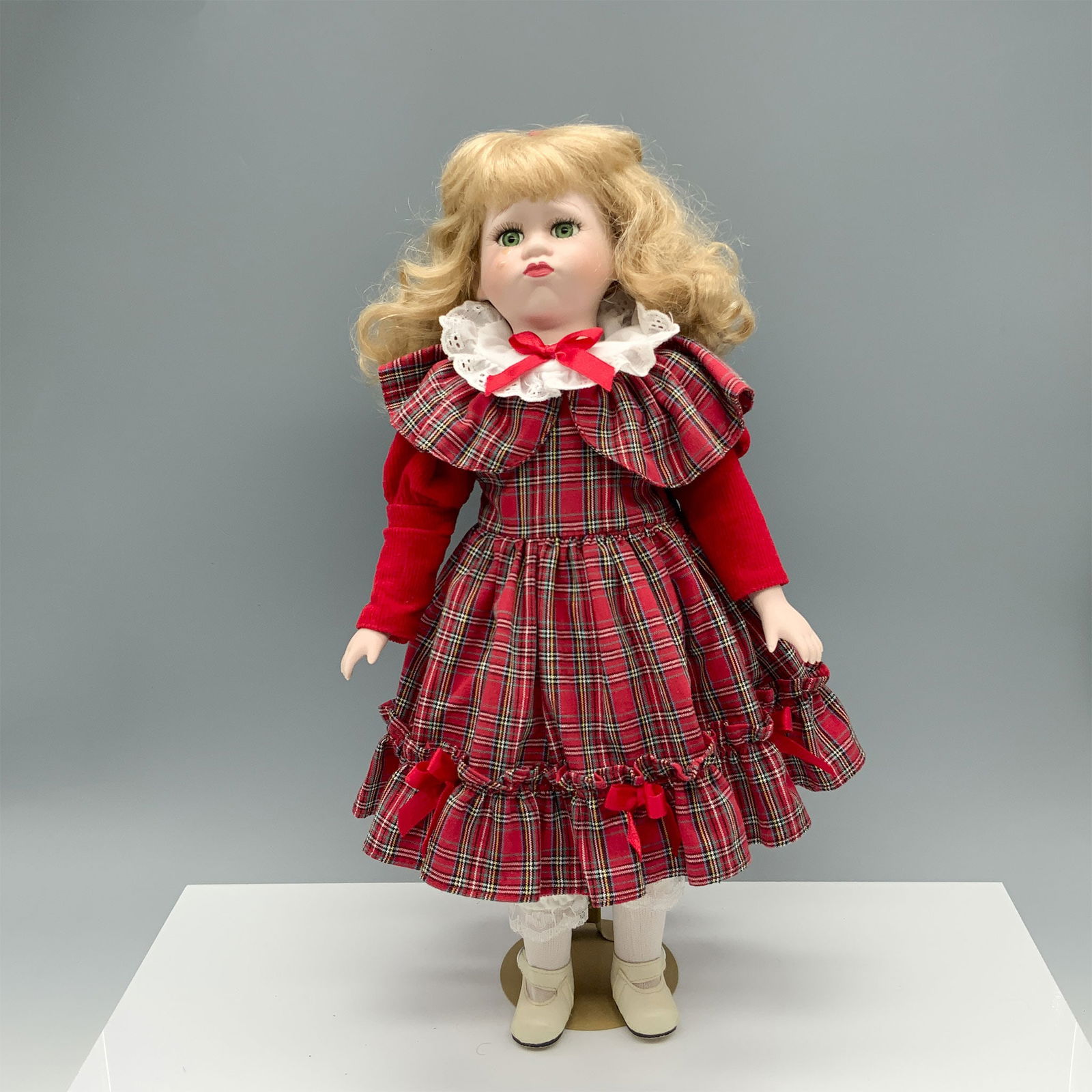 Teary Eyed Girl in a Plaid Dress Porcelain Doll: Cute green eyed blonde young girl modeling a red dress with ribboned bows on it. Metal stand included. Dimensions: 8.25"L x 7.5"W x 16"H