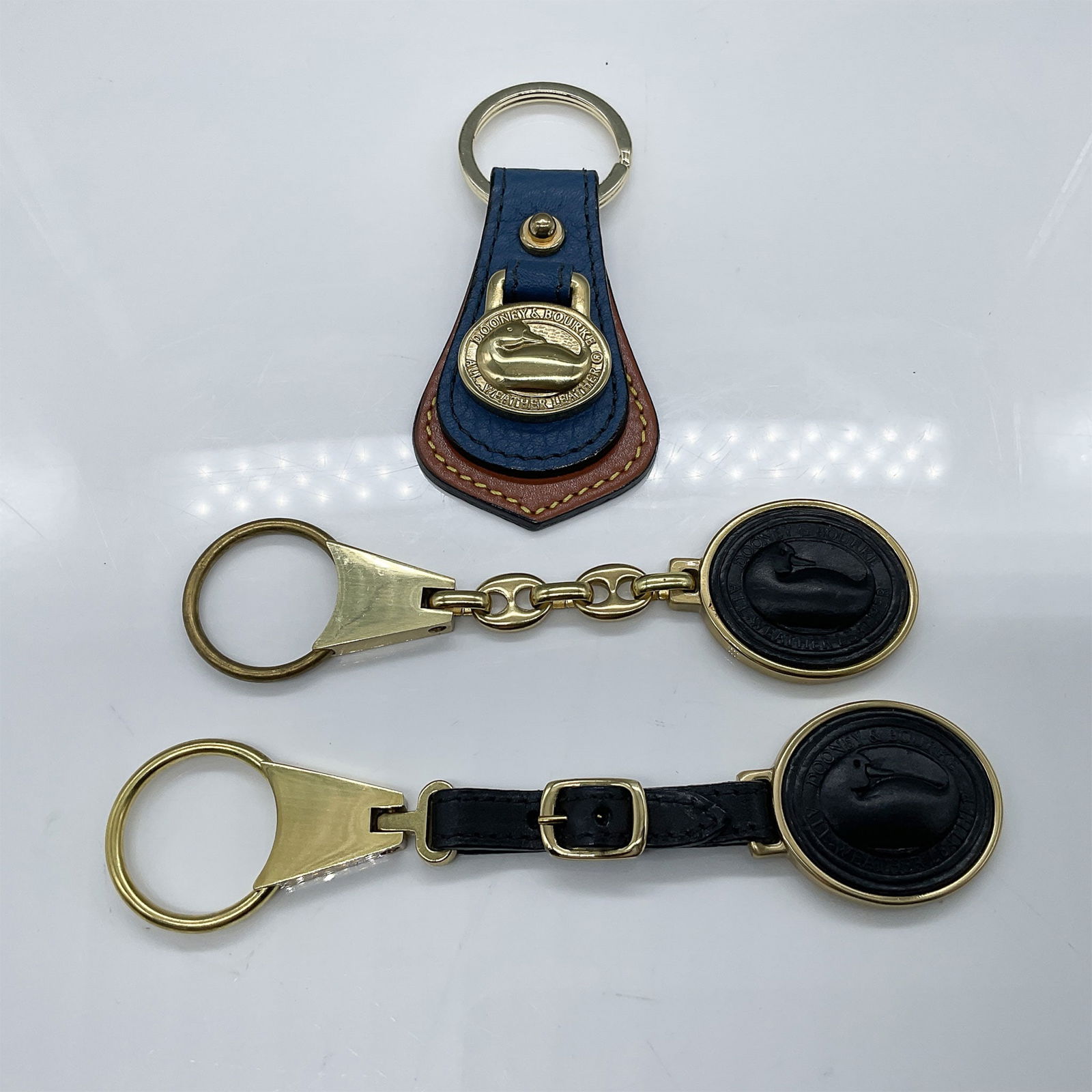 3pc Dooney & Bourke Key Chains: A 3-piece Dooney & Bourke key fob set featuring a leather and gold-tone key fob with the iconic duck logo, accompanied by two chain and belt-style key fobs. The largest piece measures 6"L x 1.5"W. Thi