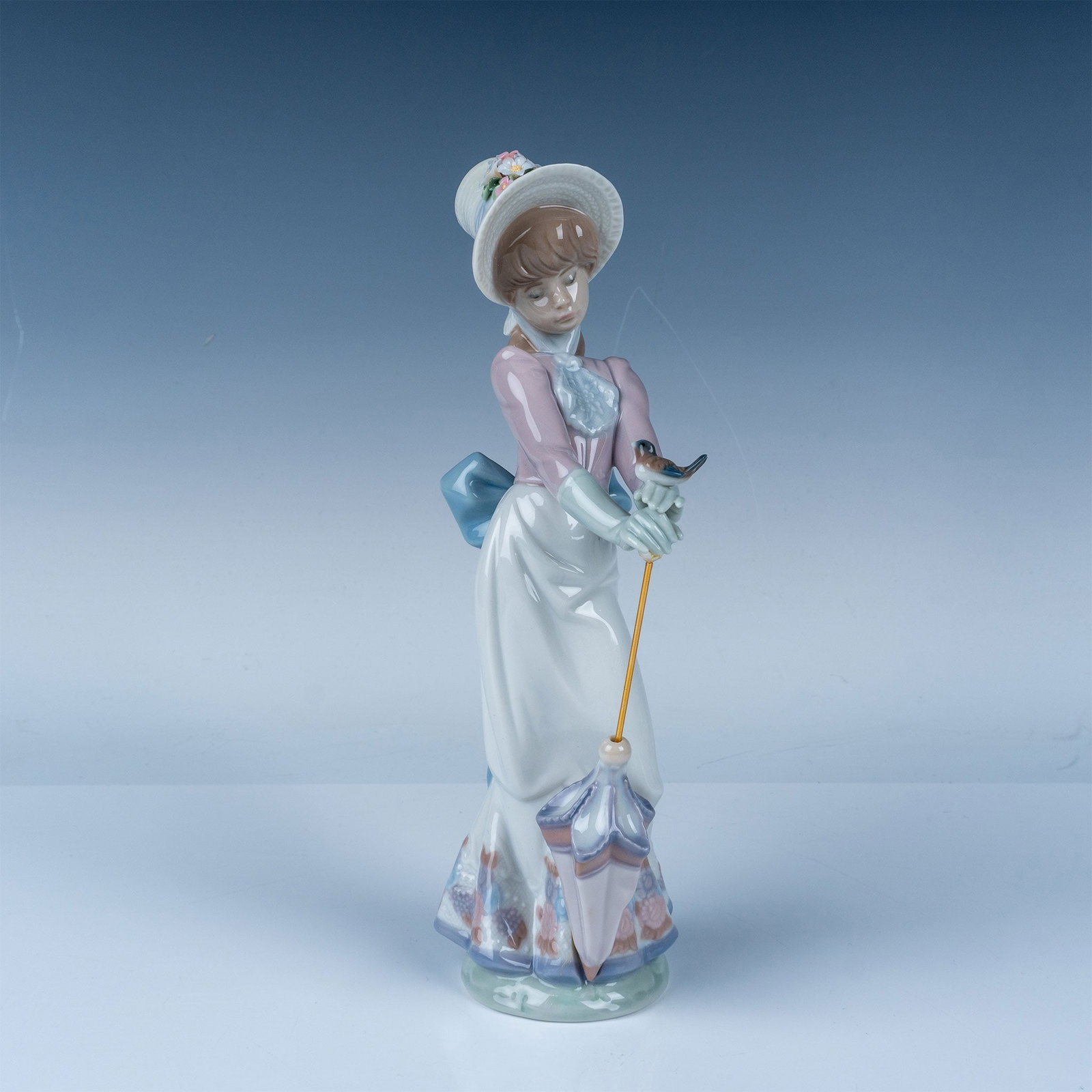 Garden Song 1007618 - Lladro Porcelain Figurine: A beautifully crafted Lladro porcelain figurine depicting a young girl holding an umbrella. Designed with soft pastel tones, the figure features hand-painted floral accents and fine details, character