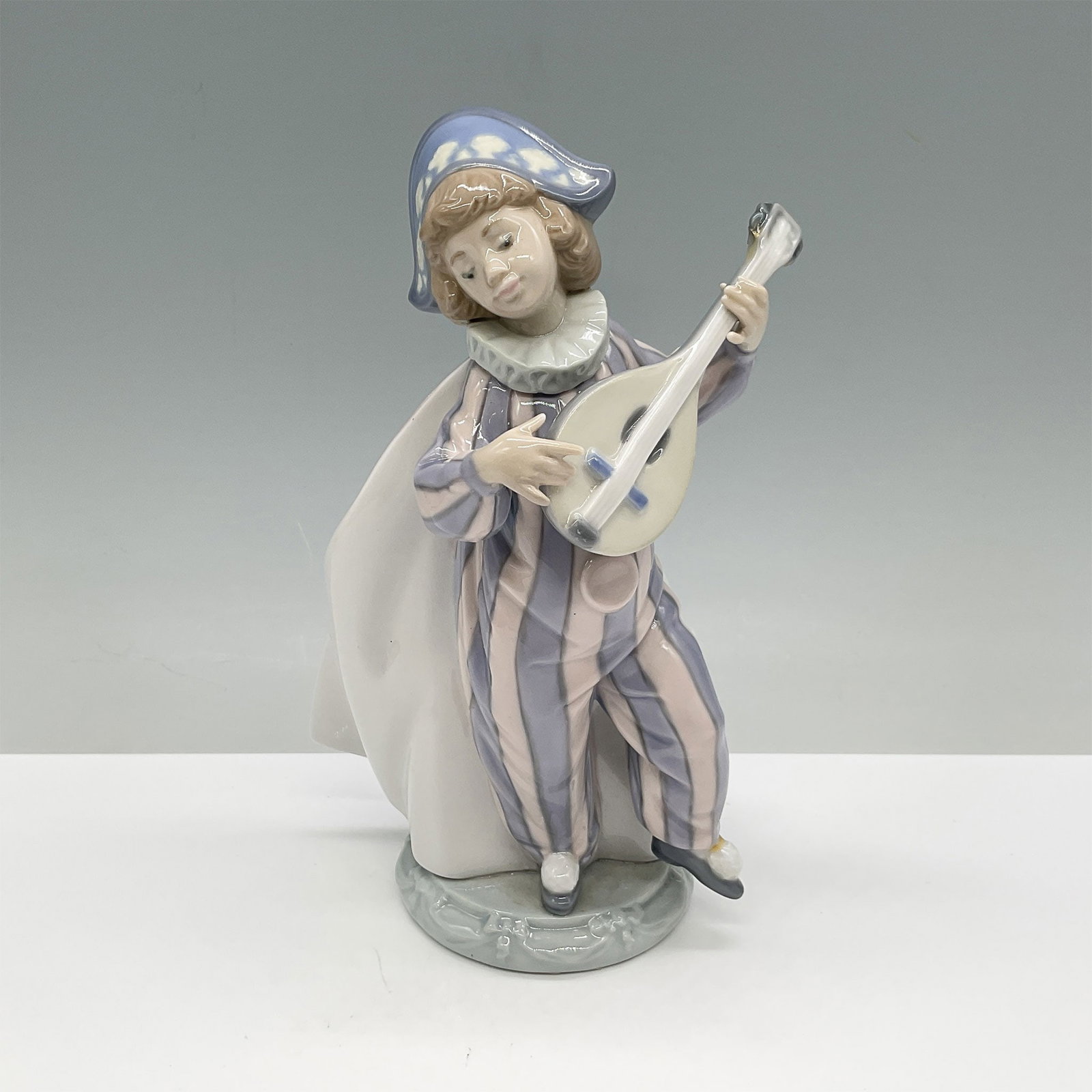 Lladro Porcelain Figurine, Mandolin Serenade 01005696: This Lladro porcelain figurine features a charming young harlequin playing a mandolin, dressed in pastel tones of pink, blue, and white with intricate hand-painted details. Designed with a whimsical f