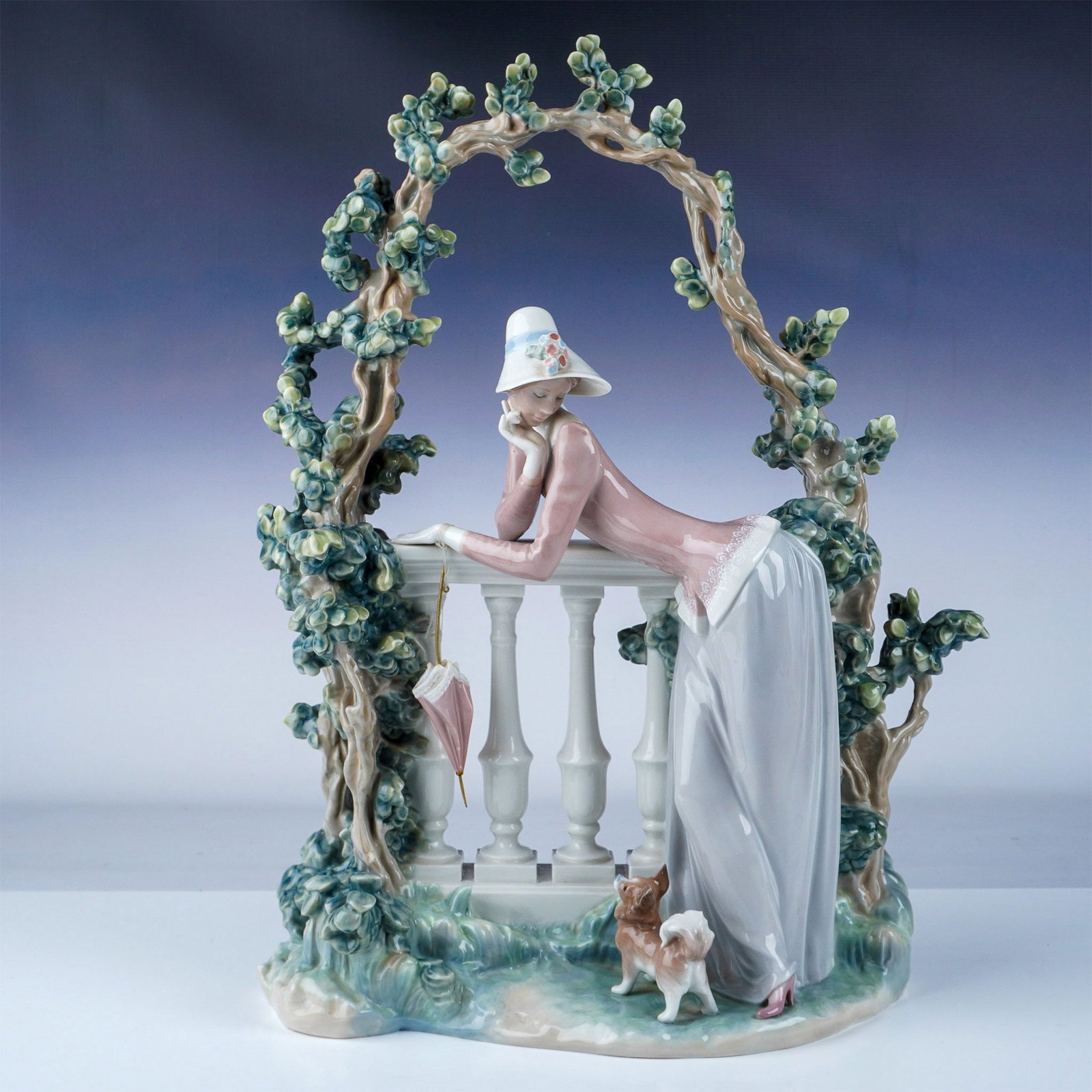 Lladro Figurine, In The Balustrade 1008680: Exquisite Lladro porcelain figurine depicting a graceful lady leaning on a balustrade beneath an arched trellis of flowering branches. Dressed in a flowing pastel gown with delicate lace details, she 