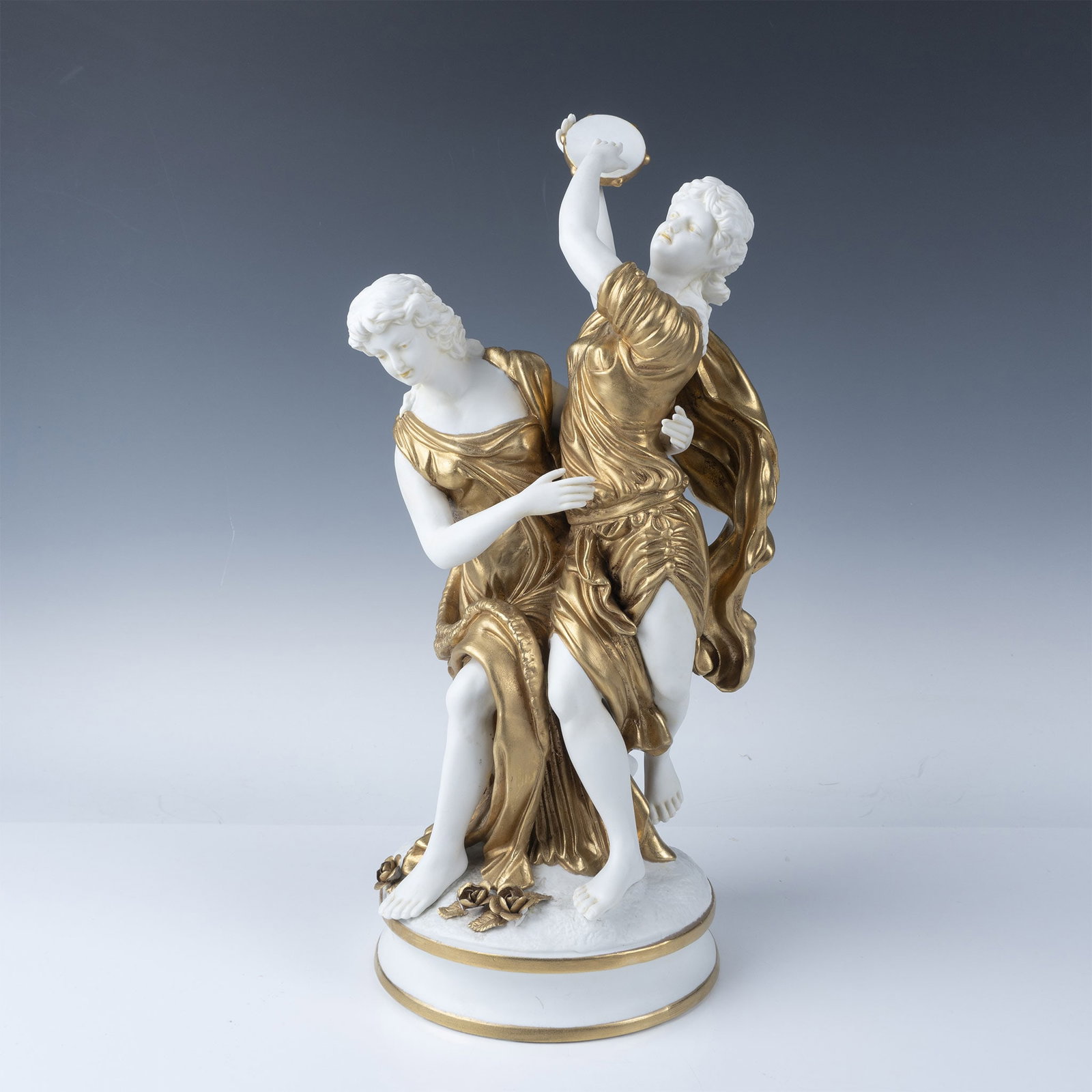 Neoclassical Porcelain Figurine, Two Classical Maidens: Featuring two elegantly draped maidens with a rich gilt and white finish. The maidens are posed gracefully, with flowing garments accented in lustrous gold toned detailing. The piece is mounted on a r