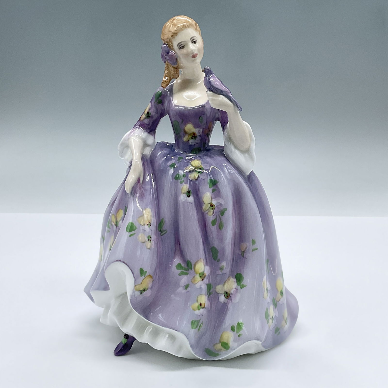 Royal Doulton Bone China Figurine, Nicola HN2839: Lovely figure of a young lady fashioned in a purple floral dress. She holds her dress with one hand, holding a bird in the other. A part of the Peggy Davies series. Royal Doulton backstamp. Artist: Pe