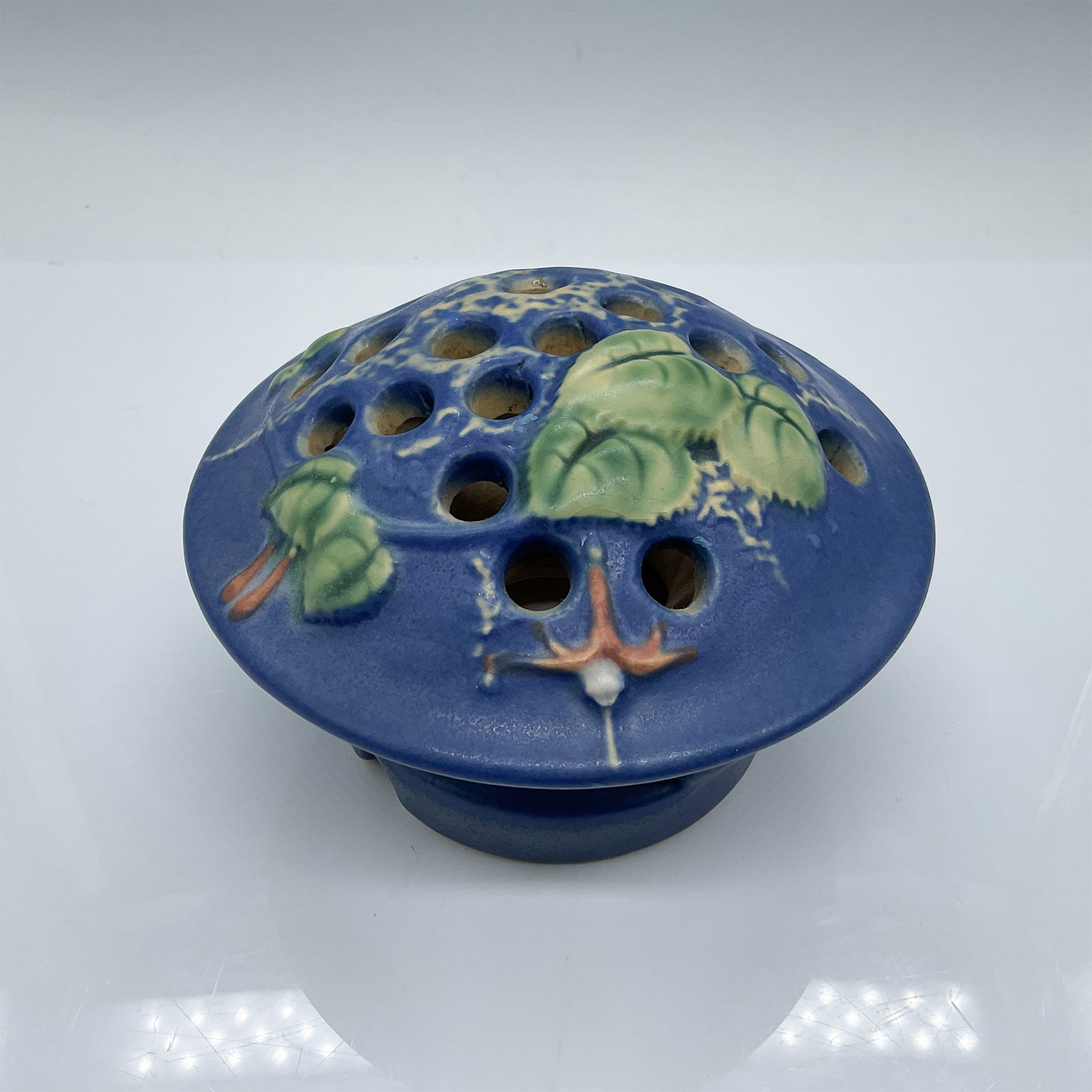 Roseville Art Pottery Clematis Flower Frog, Blue: A Roseville Art Pottery Clematis flower frog in blue, measuring 3"H x 5" in diameter. This piece features vibrant hand-painted clematis flowers with green foliage on a textured blue background. The pe