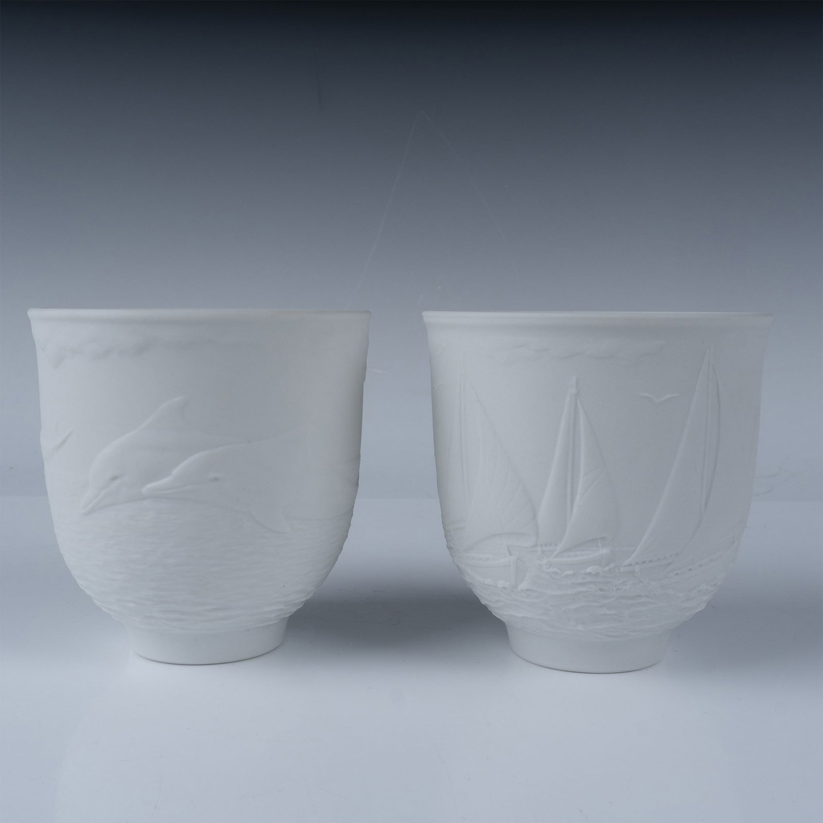 2pc Lladro Collectors Society Porcelain Cups: A pair of Lladro Collectors Society porcelain cups, featuring intricate bas-relief designs. One cup depicts playful dolphins leaping above waves, while the other portrays serene sailboats on calm wate