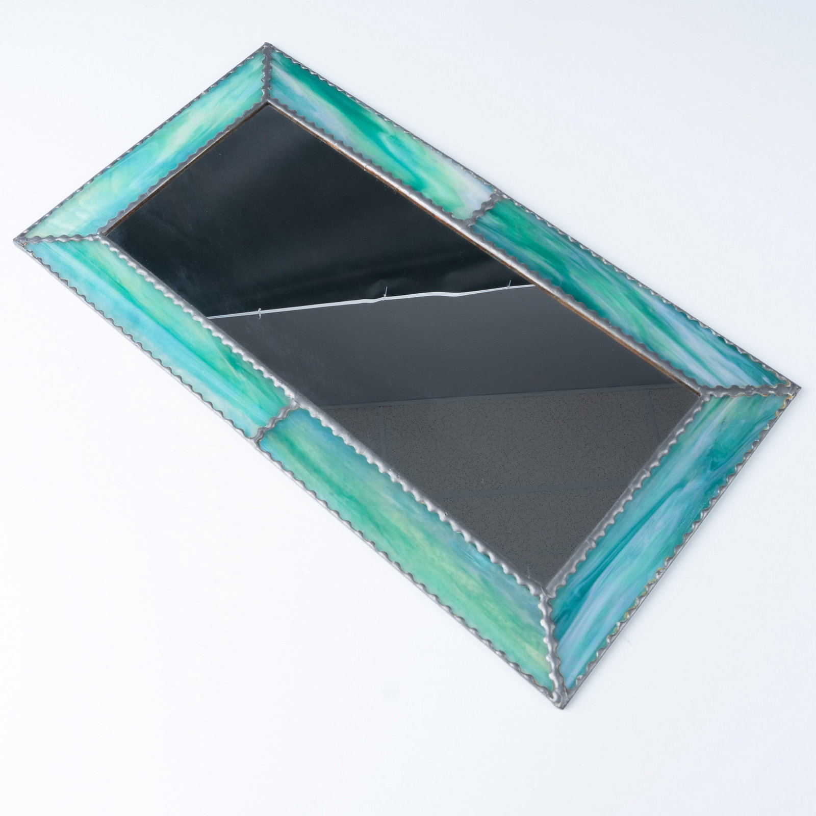 Stained Glass Framed Mirror with Iridescent Green Accents: A rectangular stained glass framed mirror featuring vibrant green and iridescent accents. The frame showcases a handcrafted design with soldered edges and a striking interplay of texture and light. Th