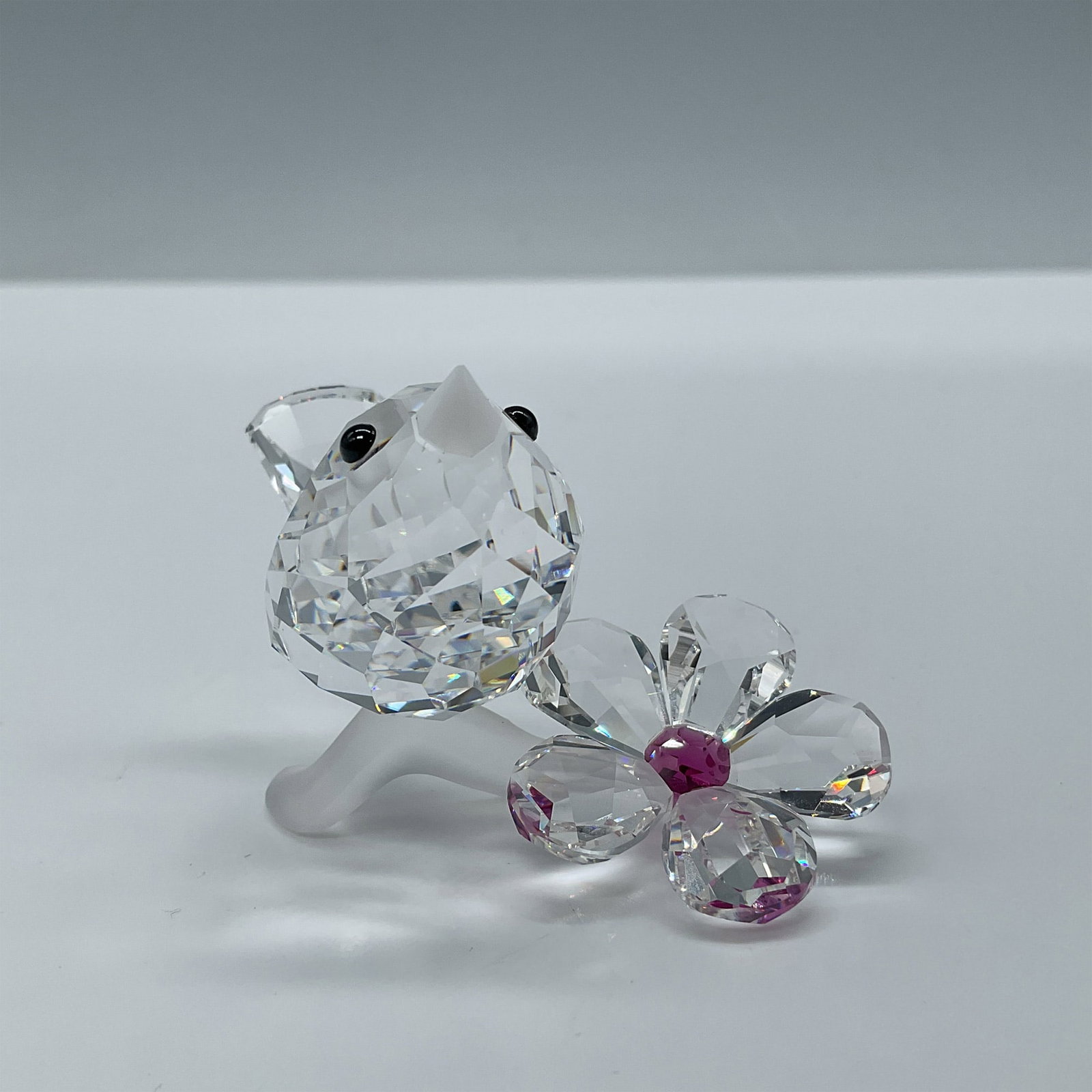 Swarovski Crystal Figurine, Baby Bird: Part of the Feathered Beauties collection. Faceted clear crystal figure has a frosted crystal beak and Jet crystal eyes sitting on satin crystal branch next to a flower with fuchsia crystal center. Sw