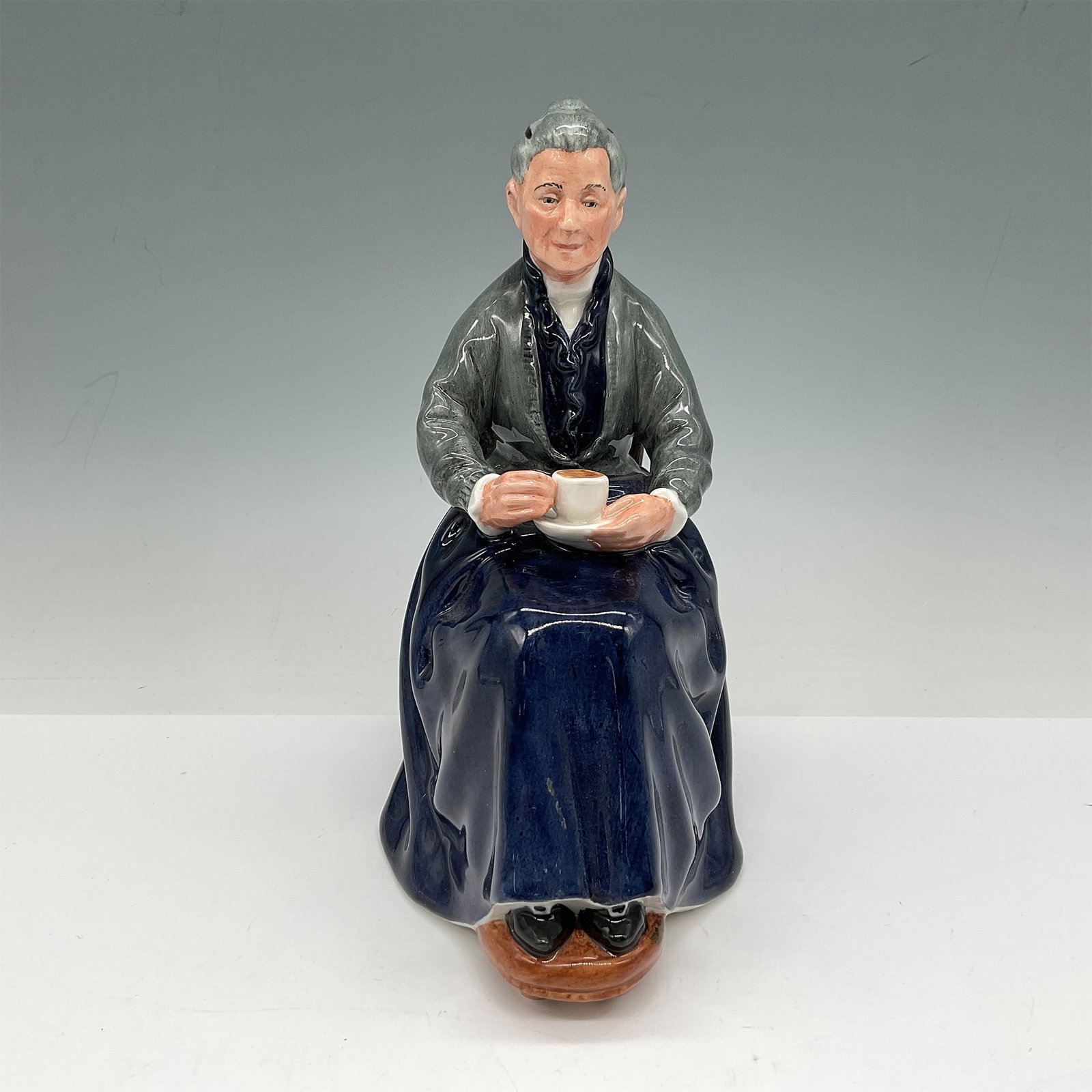Royal Doulton Porcelain Figurine, Cup of Tea HN2322: Part of the Golden Year series, figure is sitting enjoying her drink. Colored in dark blue and gray. Royal Doulton backstamp. Artist: Mary Nicoll Issued: 1964 - 1983 Dimensions: 3.5"L x 4.5"W x 7"H Co