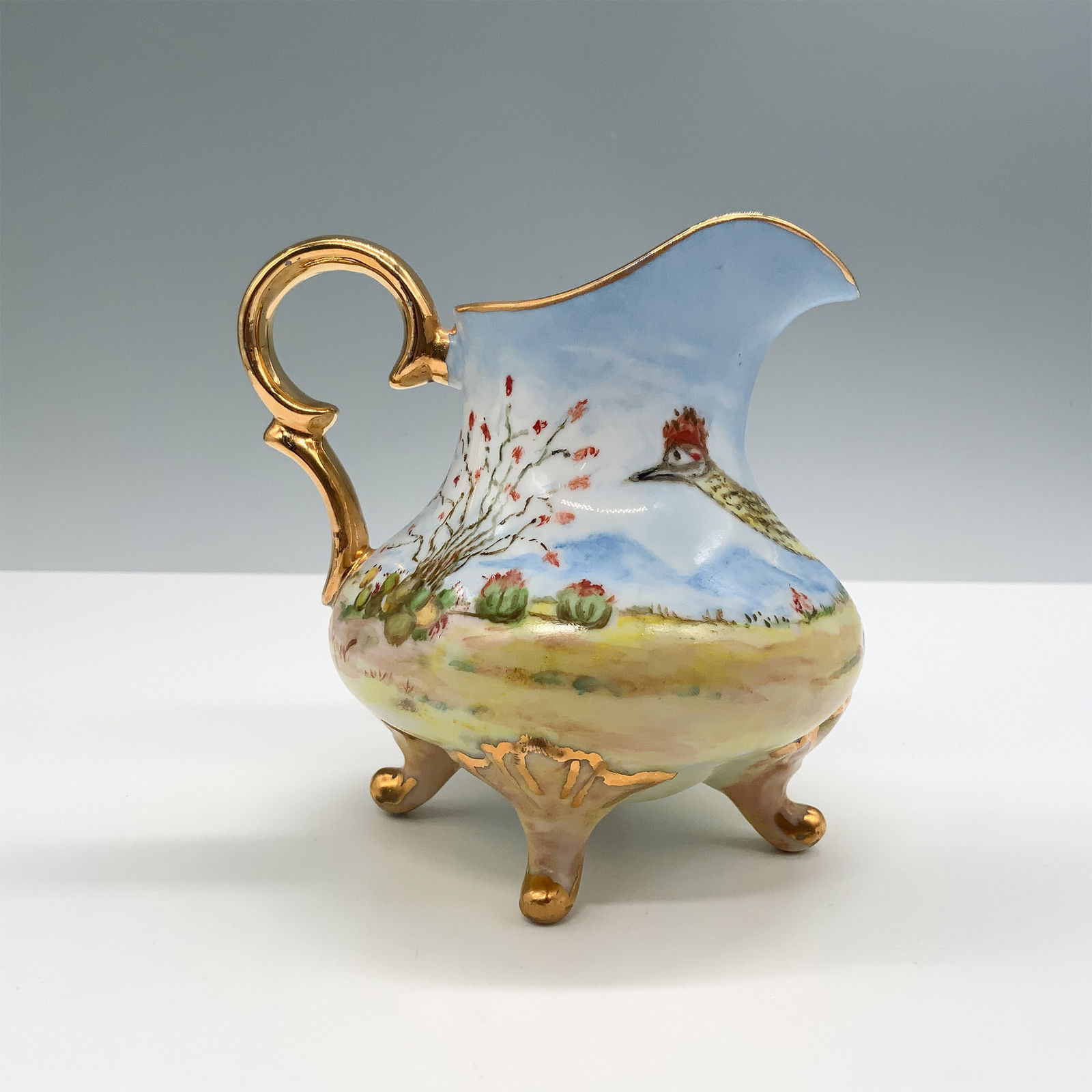 R Faulkner Hand-Painted Desert Roadrunner Porcelain Creamer: Exquisite hand-painted porcelain creamer signed by R Faulkner, featuring a vibrant desert landscape with cacti and a roadrunner on one side. The design showcases delicate brushwork on a soft blue sky