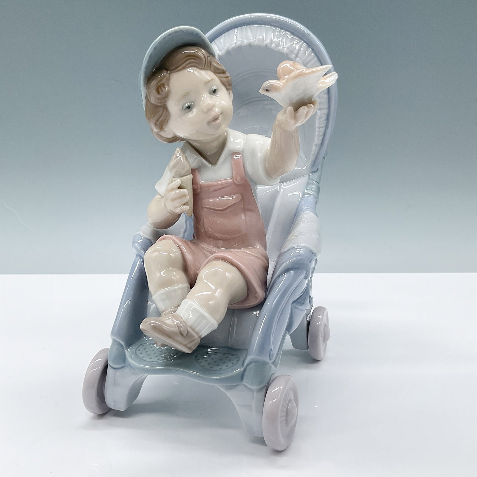 Lladro Porcelain Figurine, Bundle of Surprises 1006904: Lladro porcelain figurine titled Bundle of Surprises (06904), depicting a young child sitting in a stroller, joyfully holding a small bird in one hand and an ice cream in the other. The piece is finis