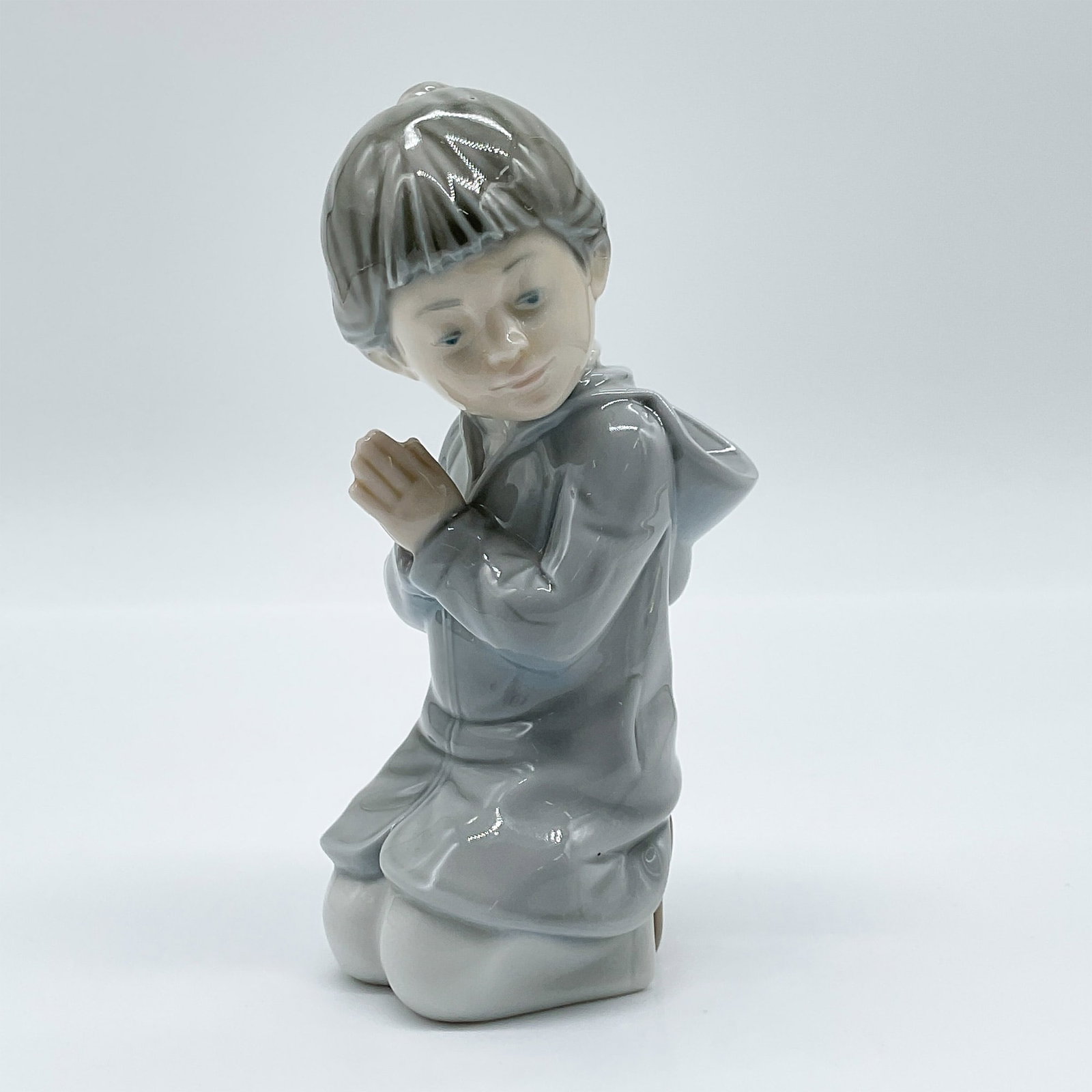 Nao by Lladro Porcelain Figurine, Guide me: This delicate Nao by Lladro porcelain figurine, titled Guide Me, captures a young girl in a moment of serene contemplation, her hands gently clasped as she kneels. Dressed in a soft gray hooded coat, 