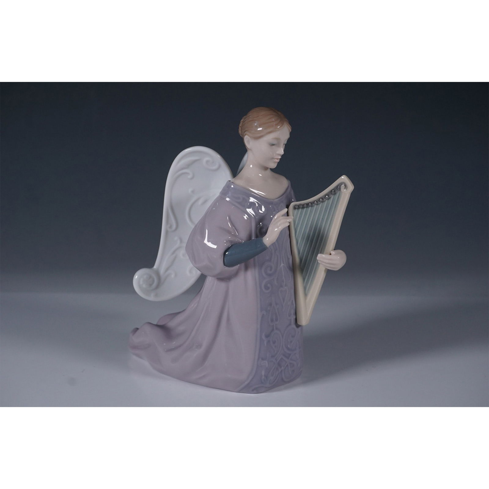 Lladro Porcelain Figurine, Harp: This stunning piece captures a serene angel kneeling while delicately playing a harp. The figurine features intricate detailing, including flowing robes in soft lavender hues, elegantly sculpted wings